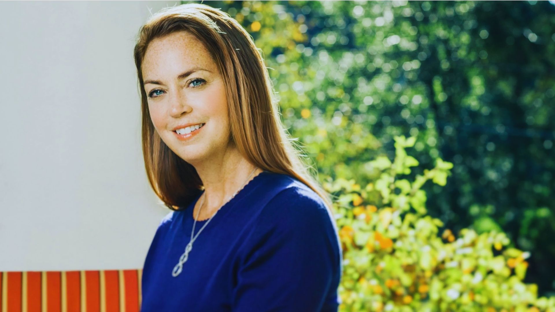 A woman wearing a blue shirt smiles warmly at the camera, exuding a friendly and approachable demeanor.