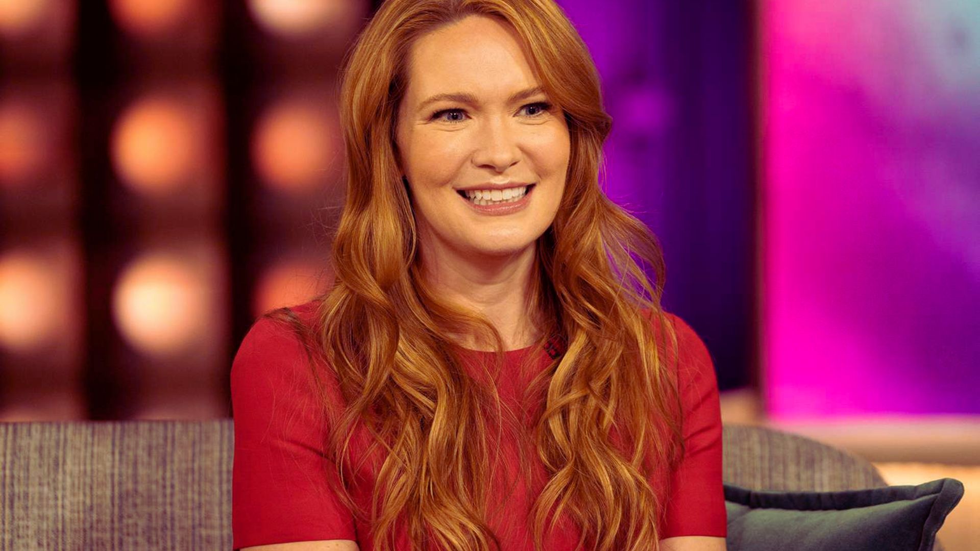 Woman with long red hair wearing a red dress sits on a sofa, smiling warmly against a softly lit purple and pink studio background.