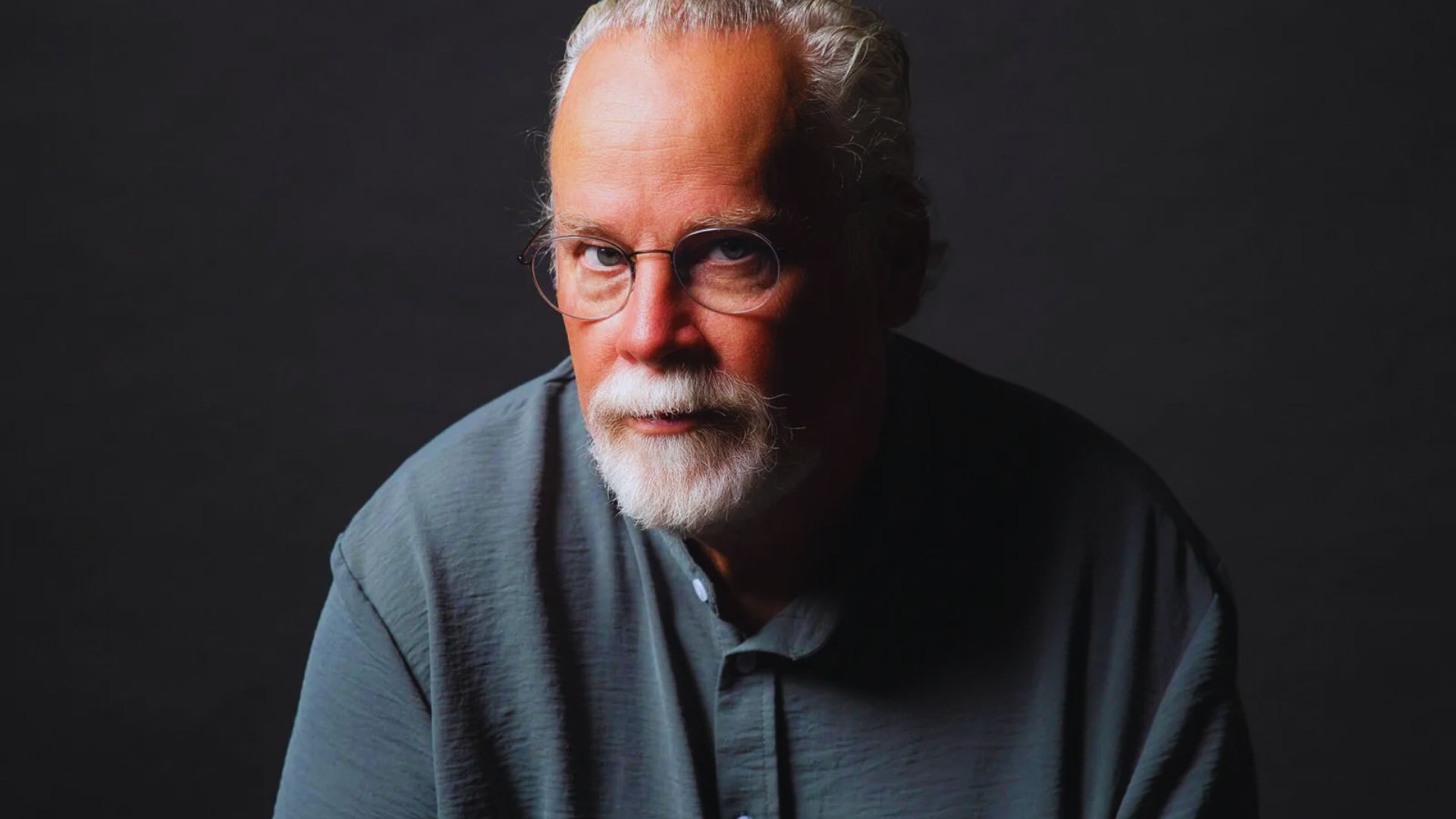 Elderly man with glasses, grey shirt, and white beard gazes thoughtfully against a dark background, conveying wisdom and introspection.