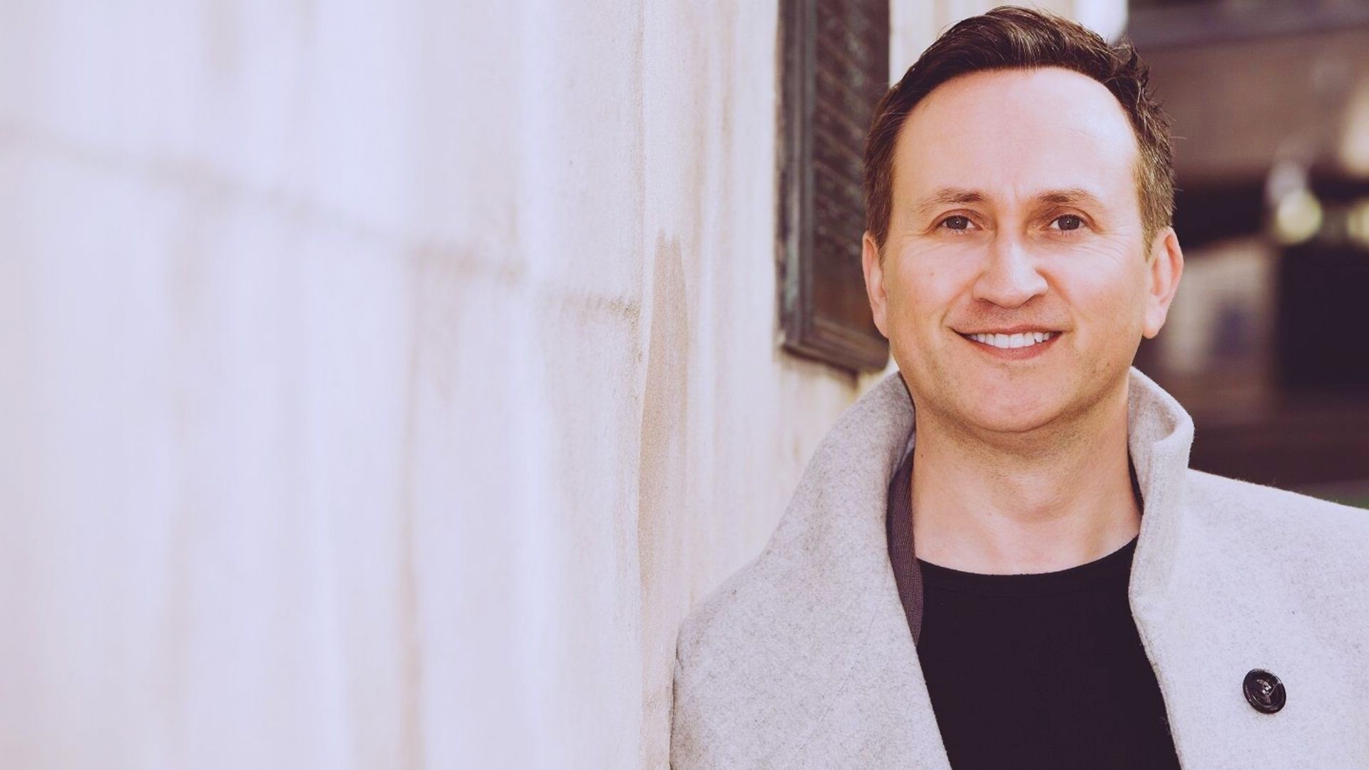 A man in a grey coat stands beside a textured wall, looking thoughtfully into the distance.