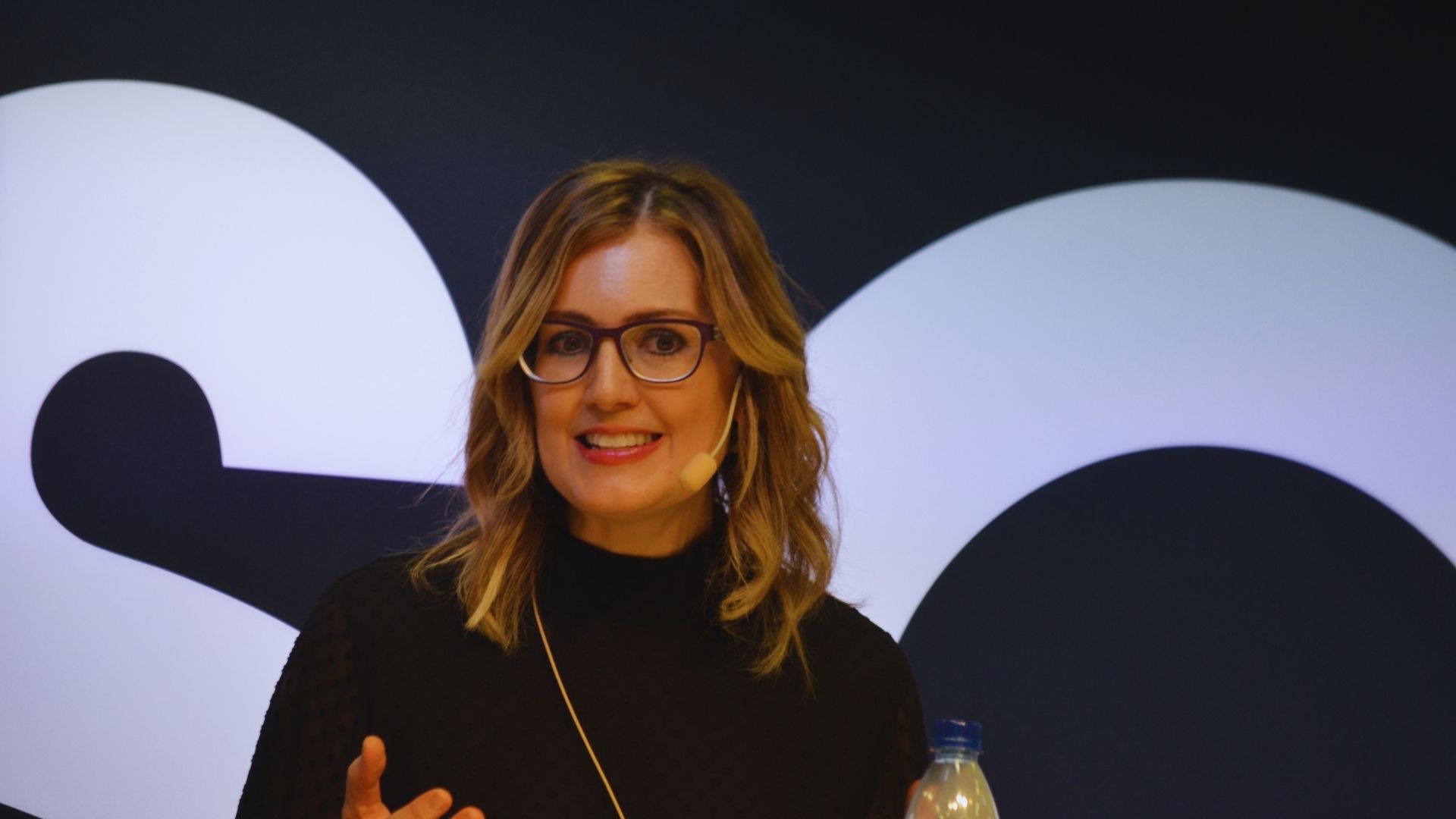 A woman with glasses, wearing a black top, speaks on stage with a headset mic. She appears confident against a backdrop of large, bold, partially visible letters.