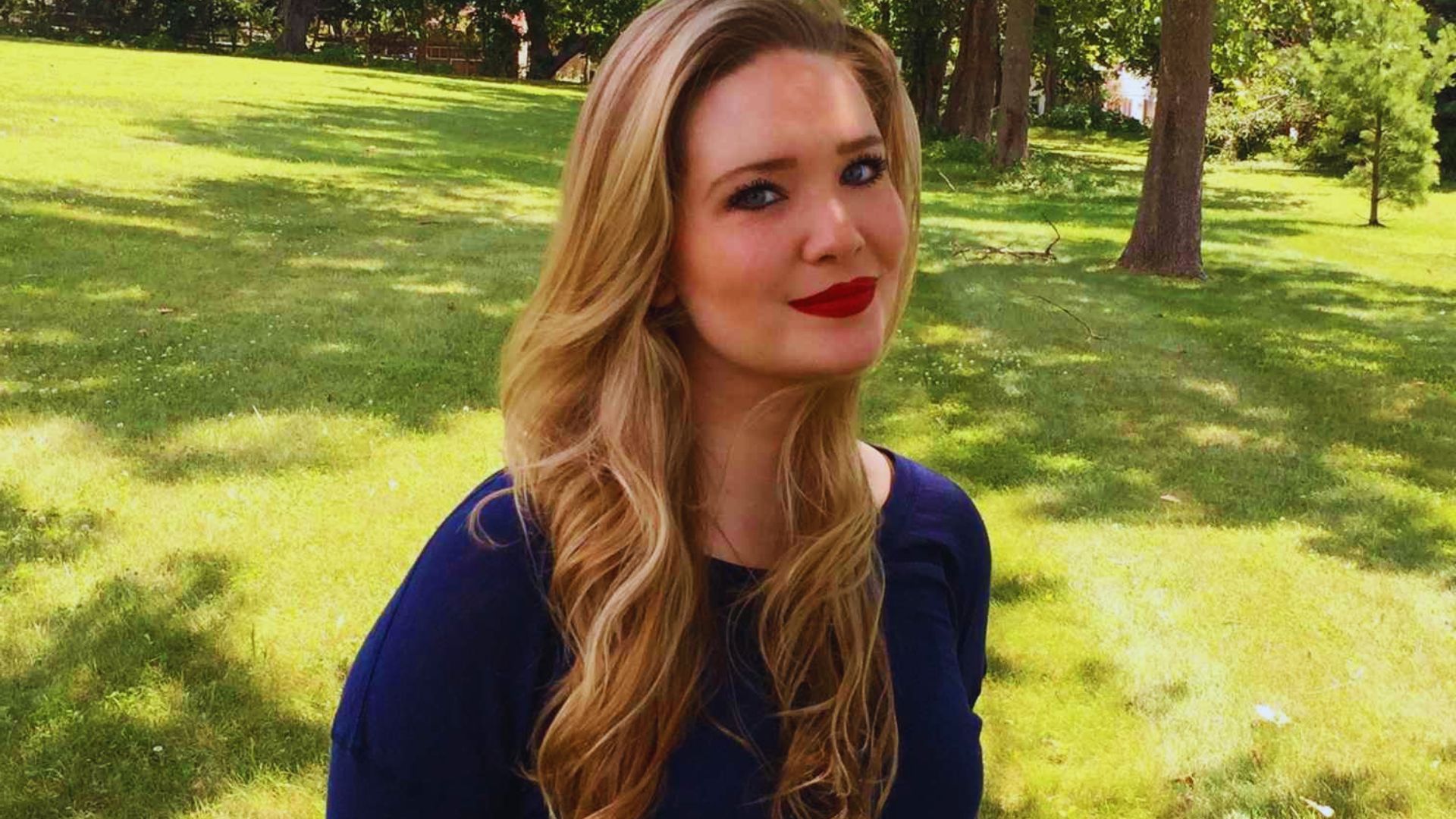 A woman with long blonde hair and red lipstick smiles confidently at the camera.