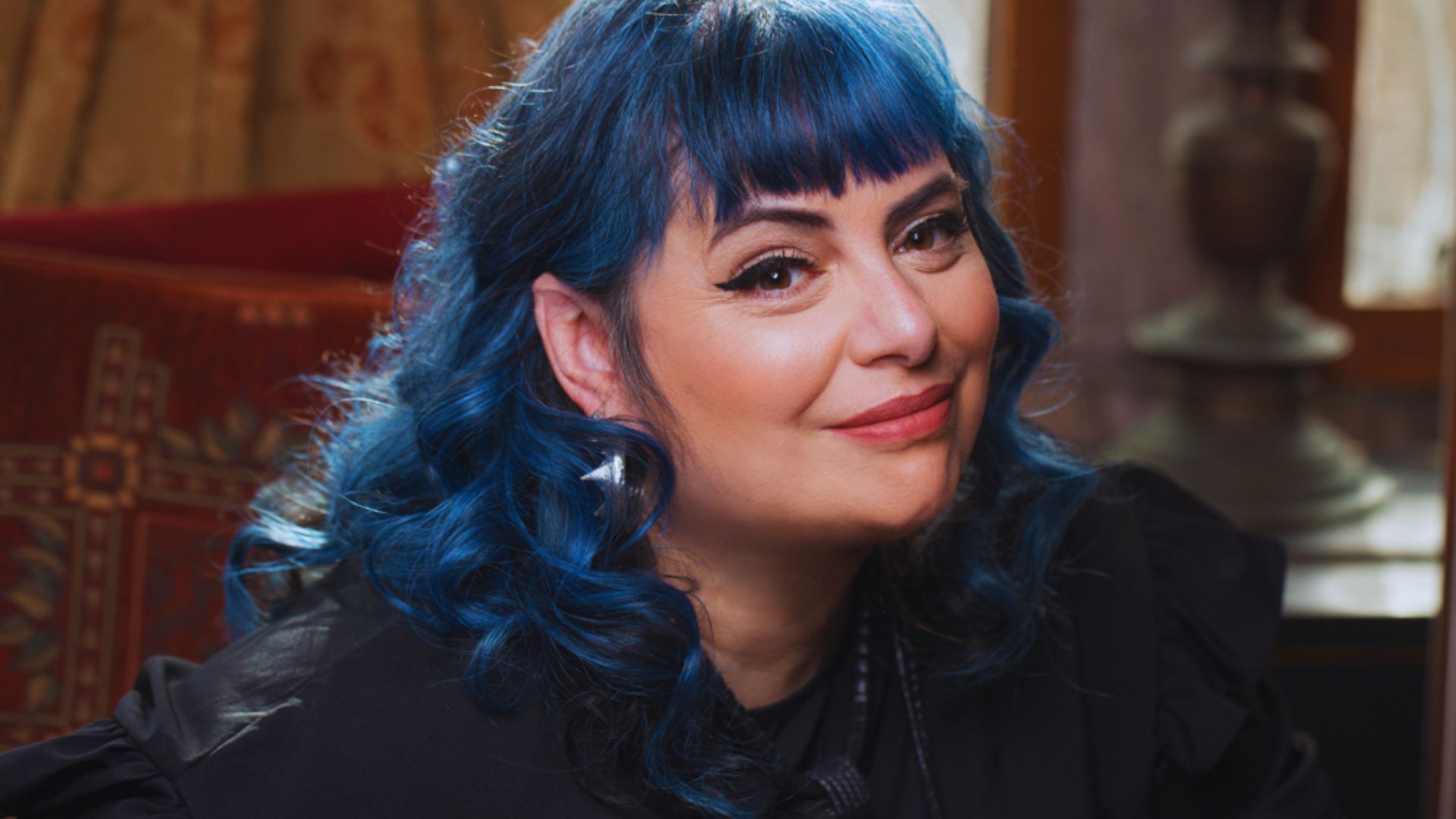 Woman with vibrant blue hair and a warm smile poses indoors. The setting is cozy, featuring patterned textiles and soft lighting, conveying a welcoming atmosphere.