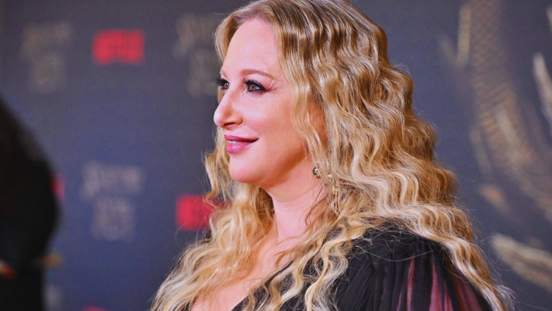 Blonde woman with wavy hair, wearing a black dress, smiles at a red carpet event. The background is dark with patterned details, evoking elegance.