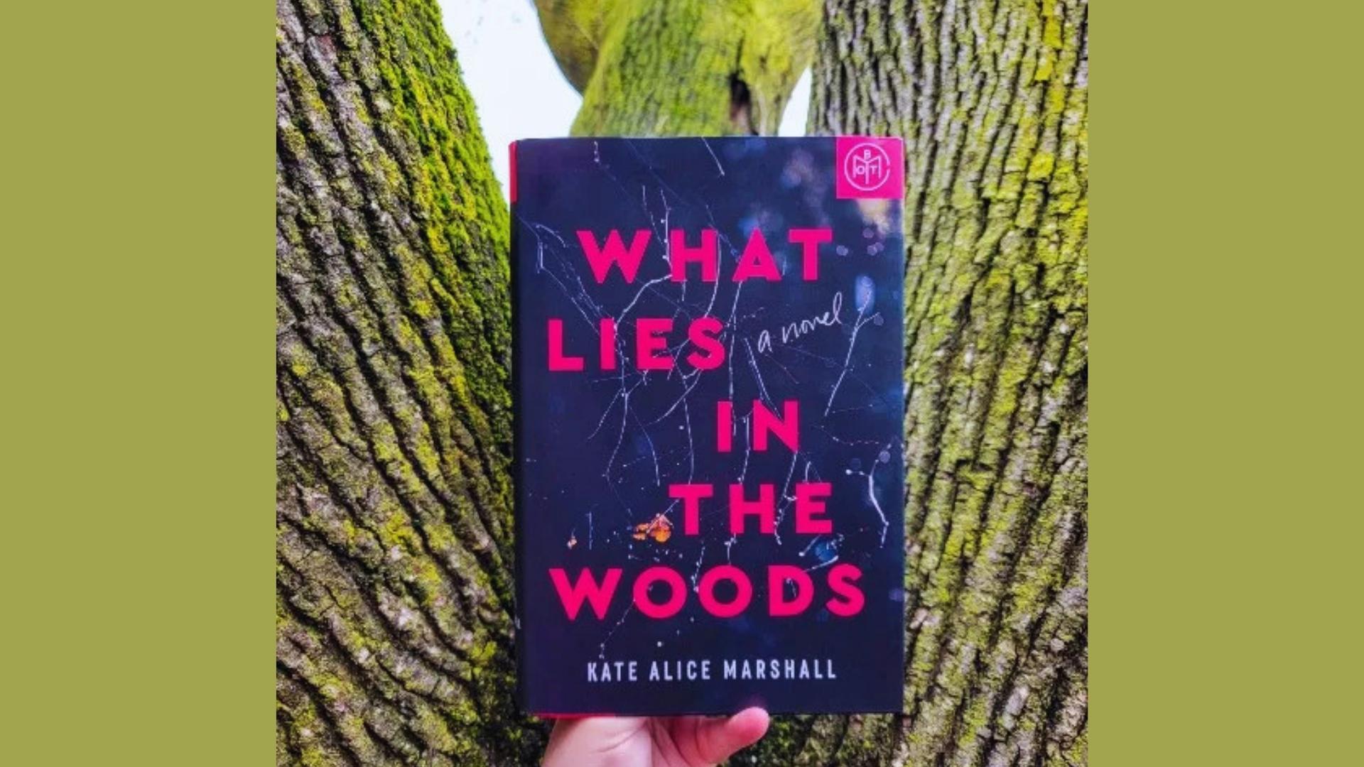 What Lies in the Woods by Kate Alice Marshall hardcover held up against mossy tree trunks with Book of the Month club sticker