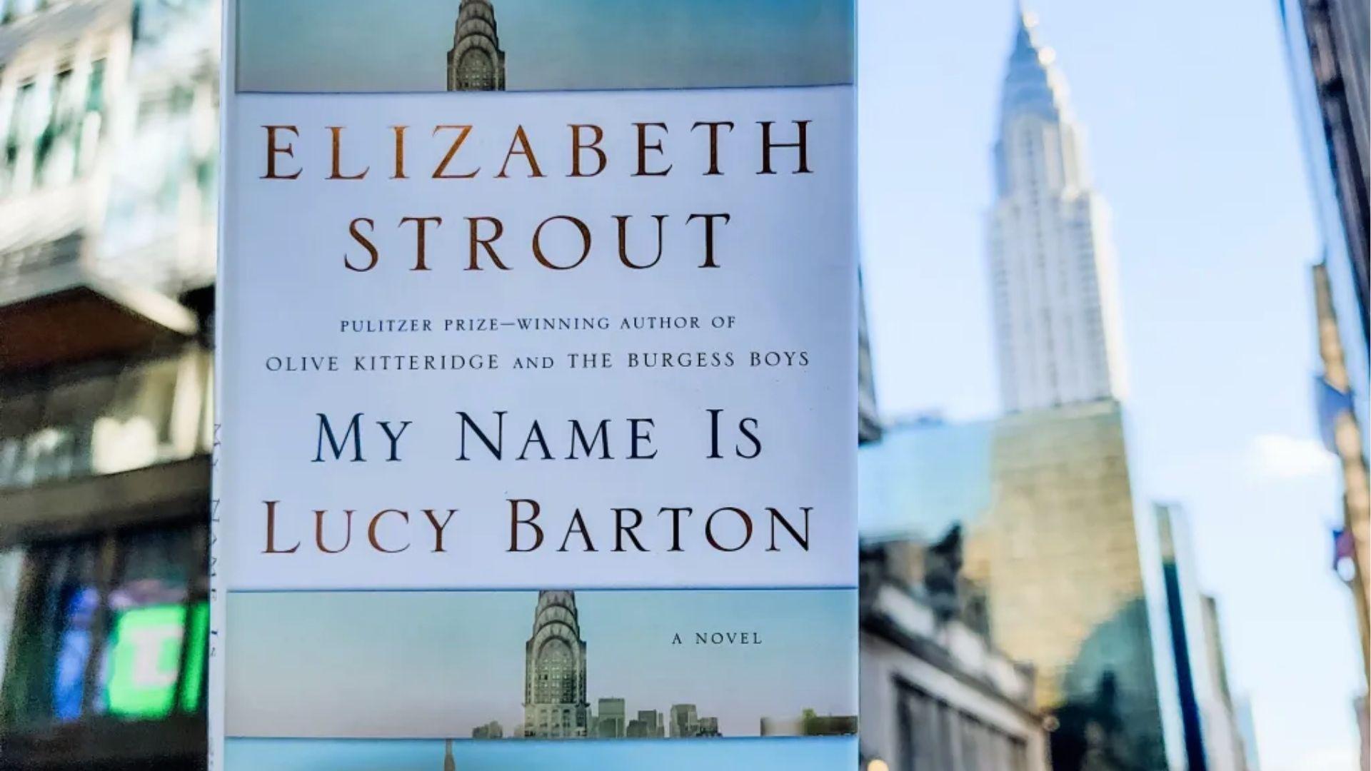 Close-up of My Name Is Lucy Barton held up against a blurred New York City street, with the Chrysler Building visible behind it.