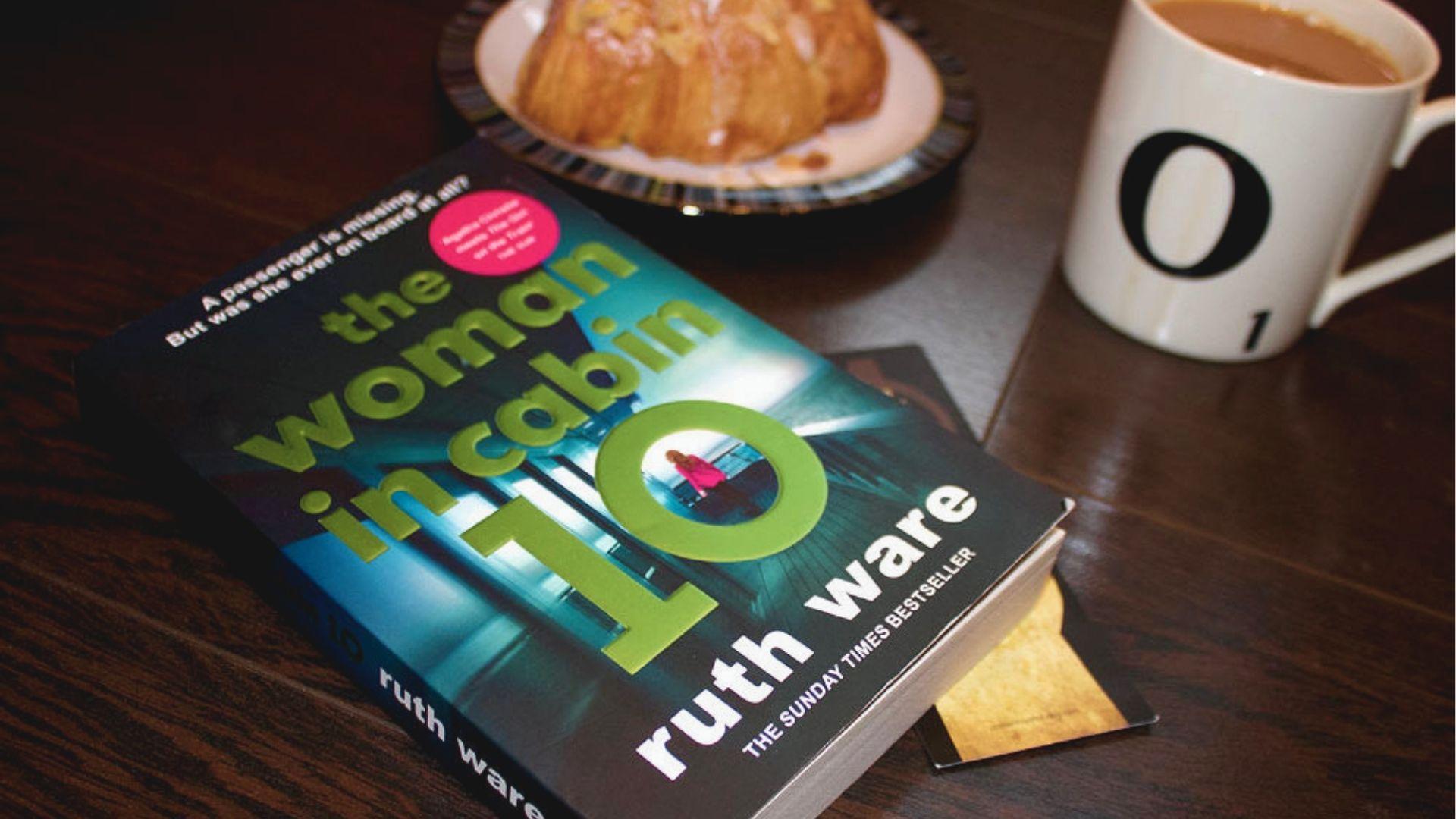 Paperback copy of The Woman in Cabin 10 lying on a dark wood table beside a croissant and coffee mug.