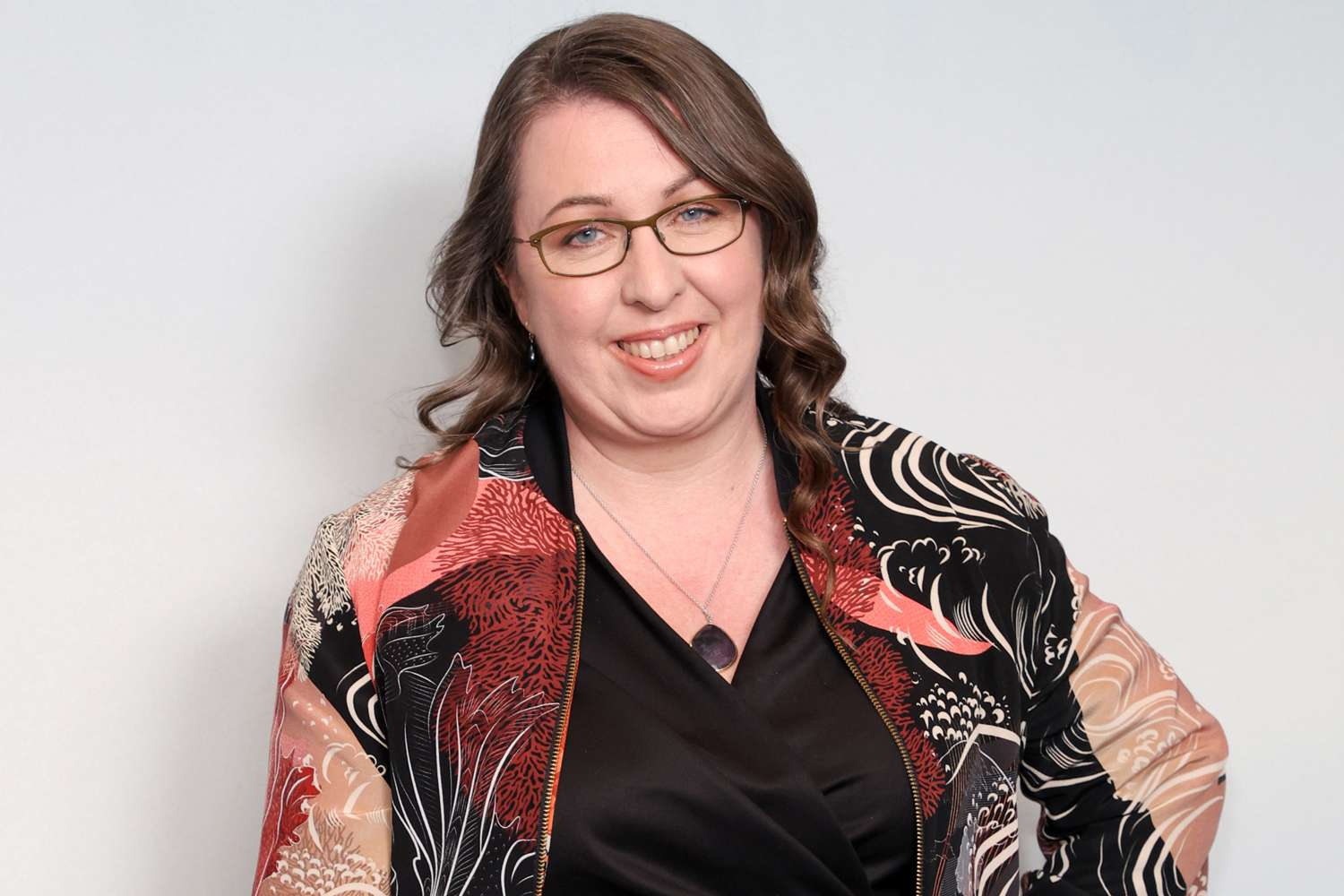 Headshot of a smiling woman with glasses wearing a patterned jacket over a black top against a plain backdrop.