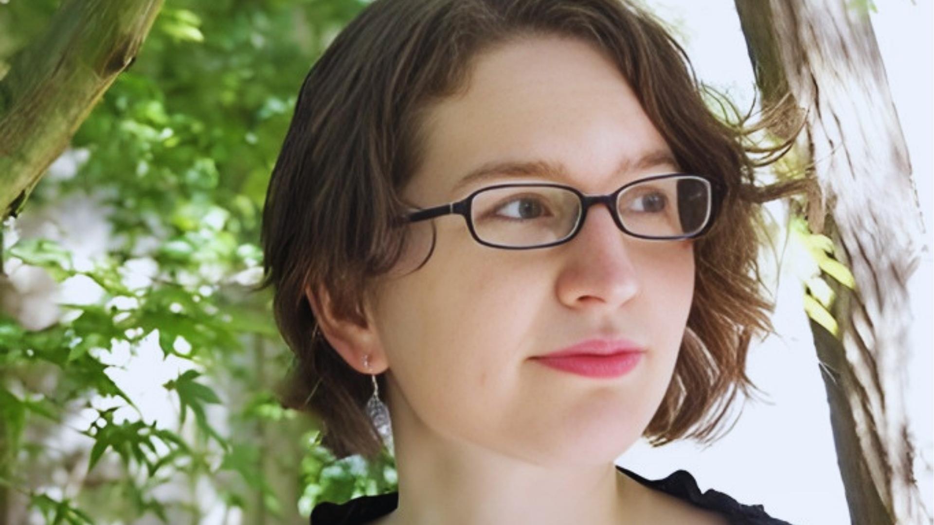 Author photo of Kate Alice Marshall wearing glasses and a black top, standing outdoors among green leaves and trees