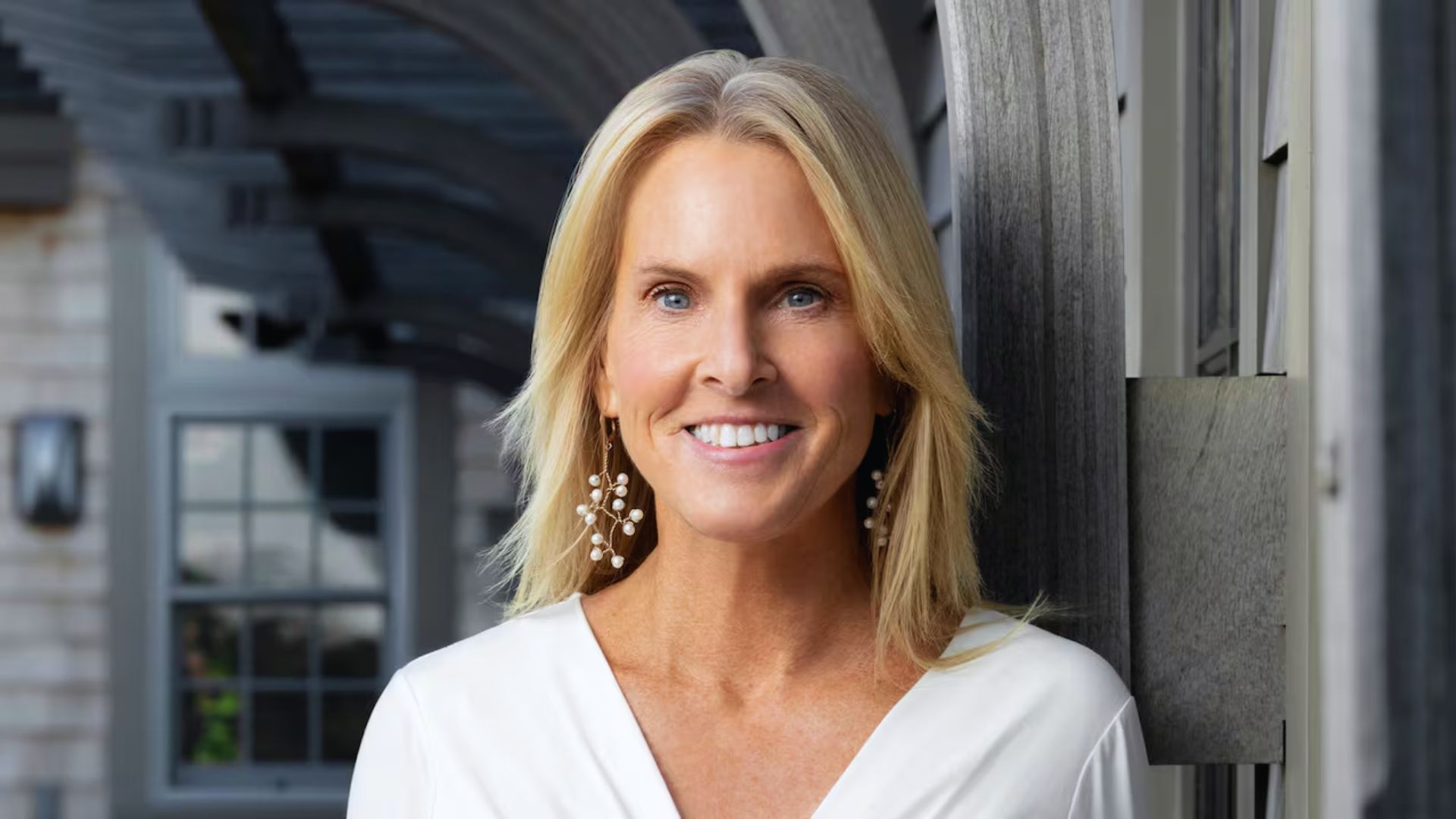 Elin Hilderbrand, author of "The Perfect Couple," stands outside with blonde hair and earrings, smiling at the camera.