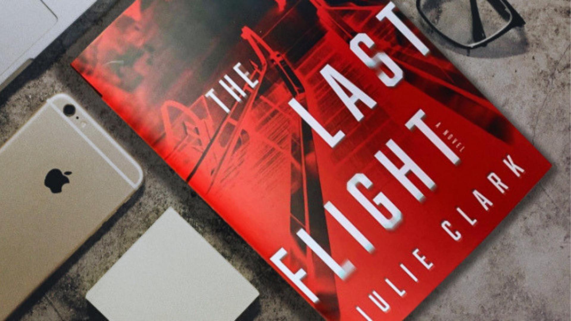 Flat lay of The Last Flight by Julie Clark paperback on desk next to iPhone, glasses, and notepad