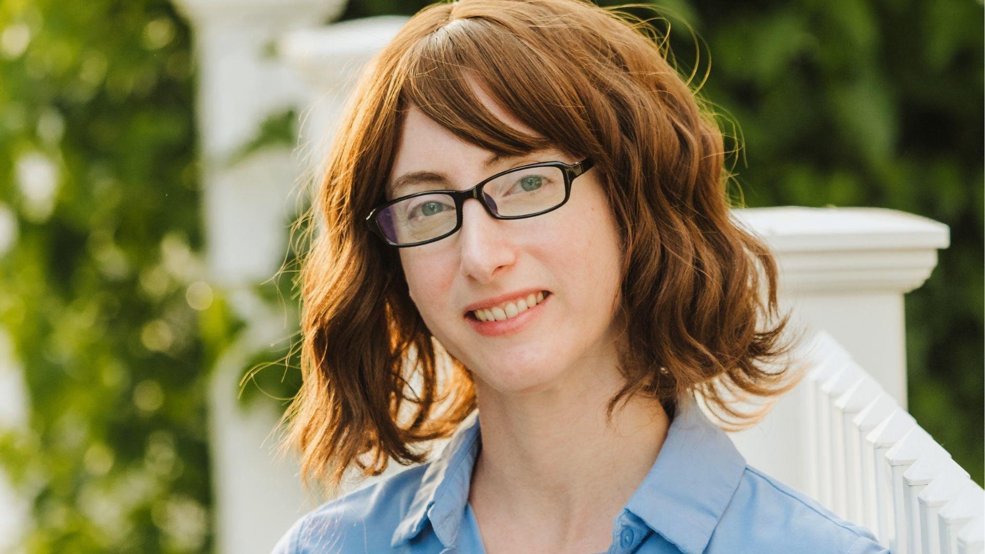 Author Freida McFadden smiling outdoors by a white fence, wearing glasses and a light blue shirt.