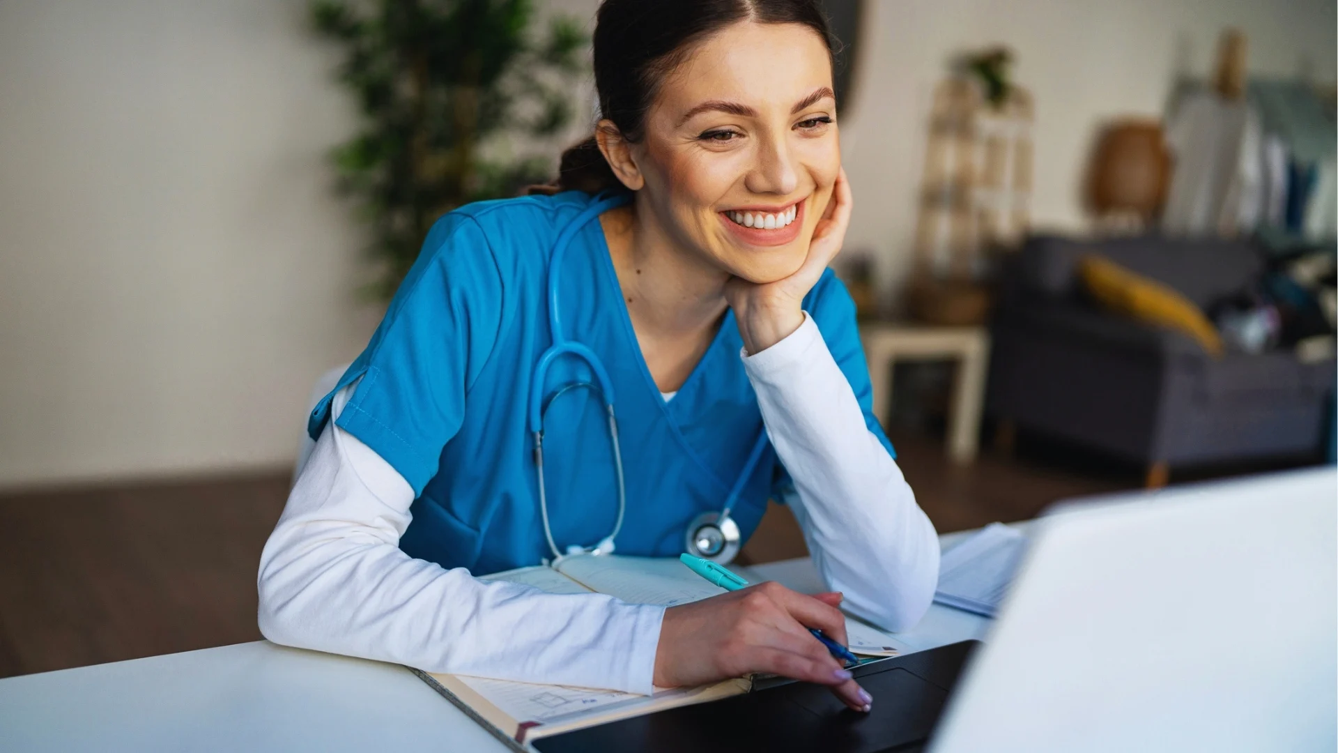 Nursing student in blue scrubs with stethoscope studying on laptop at desk with notebooks and papers.
