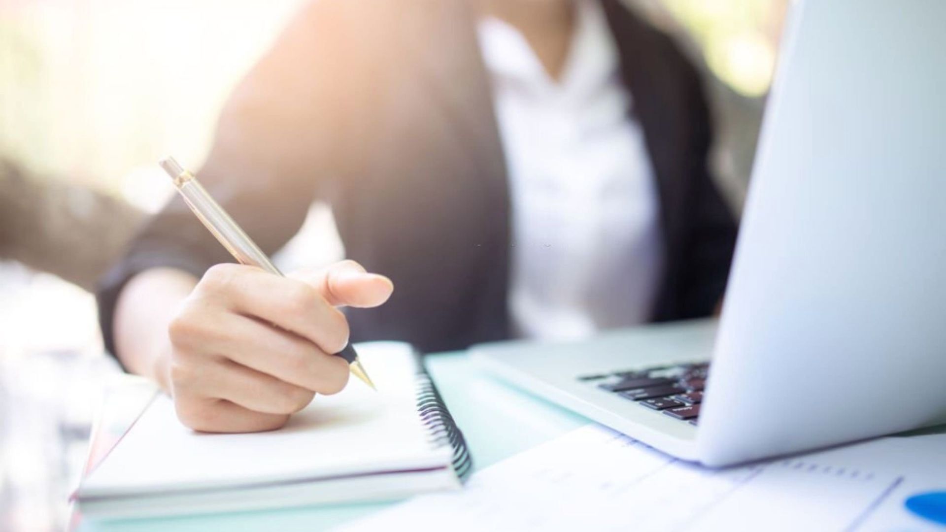 Businesswoman writing in a notebook with a pen, while a laptop is open in front of her on a desk.