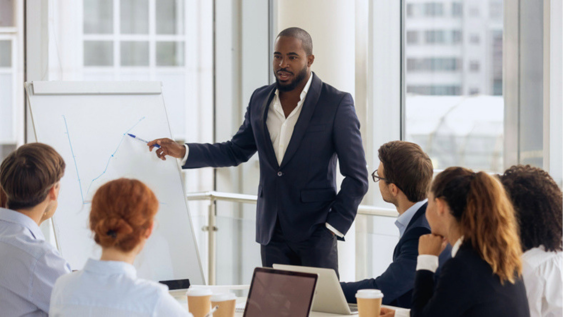 Business presenter in dark suit explaining line graph on whiteboard to team during corporate meeting