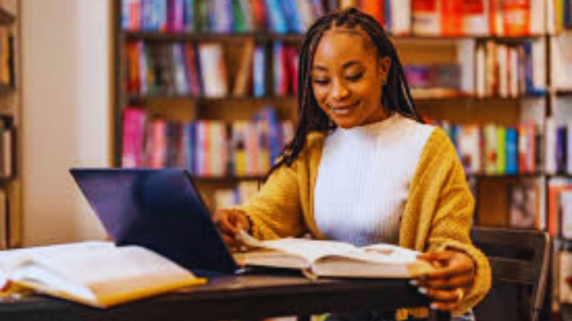 A student studying in a library with a laptop and an open book, focused on their work.