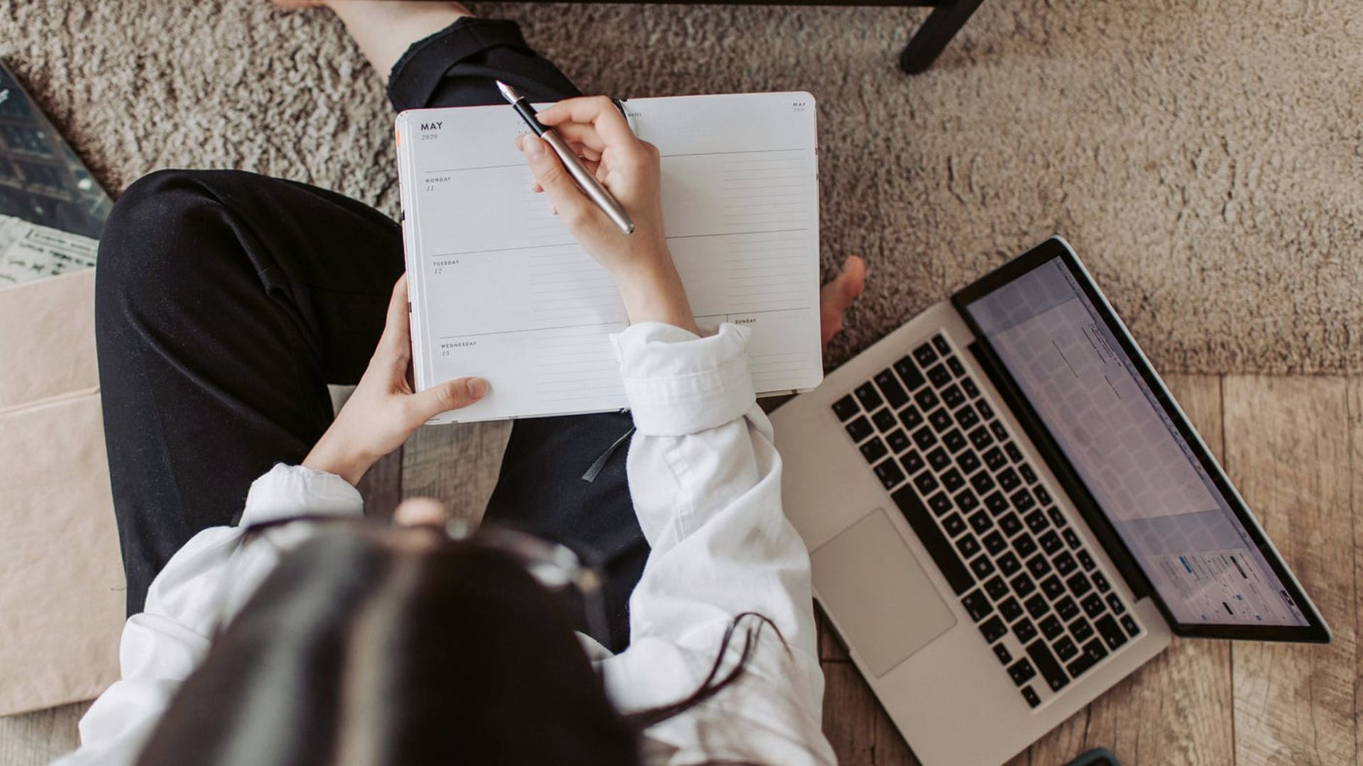 A woman in a business suit sits on the floor, working on a laptop with a notebook beside her.