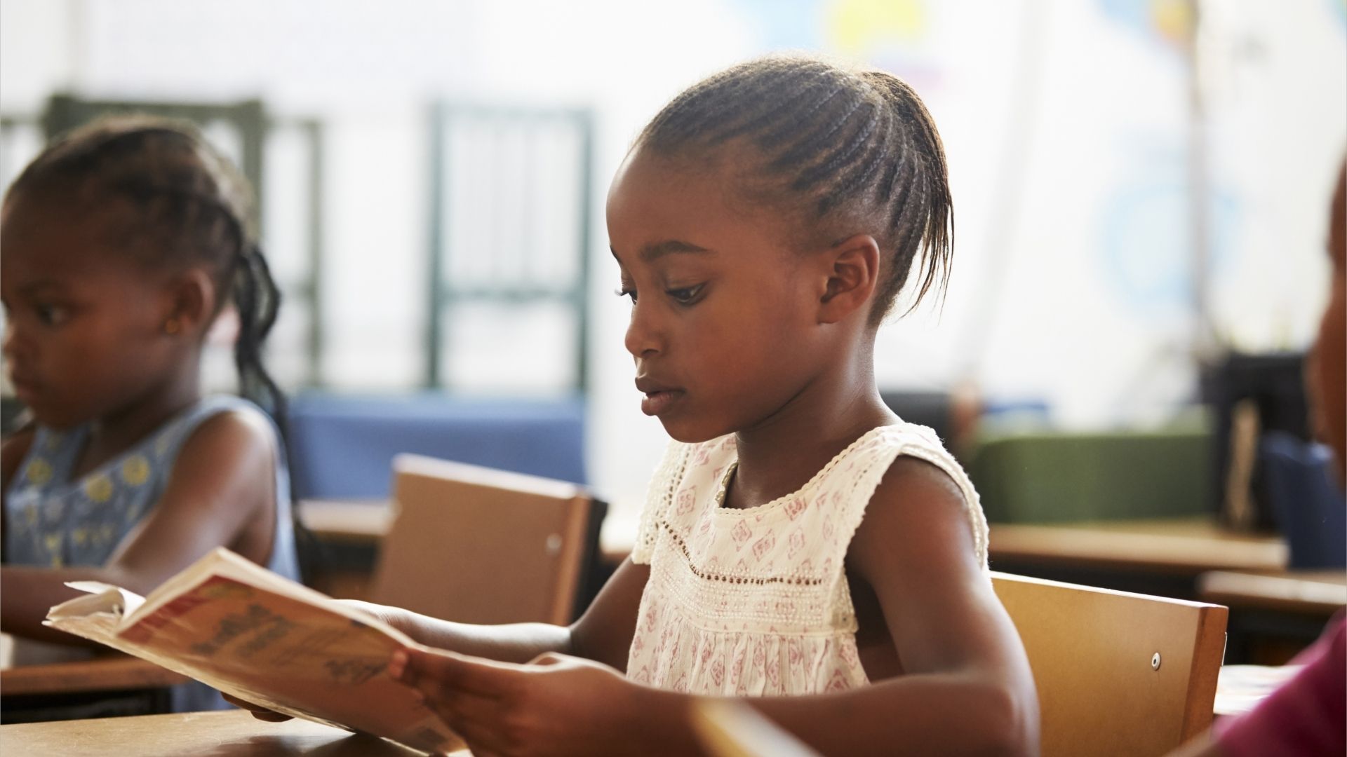 A young girl sits at a desk in a classroom, deeply focused on reading a book.