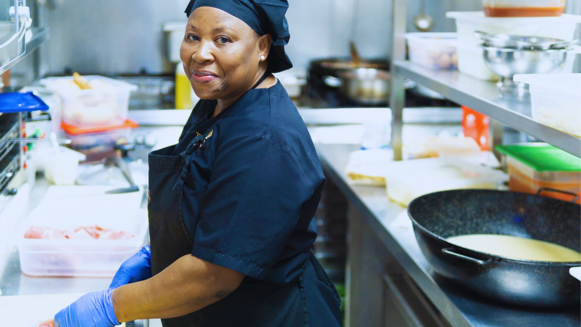 A woman dressed in black cooking in the kitchen, smiling while looking at the camera.