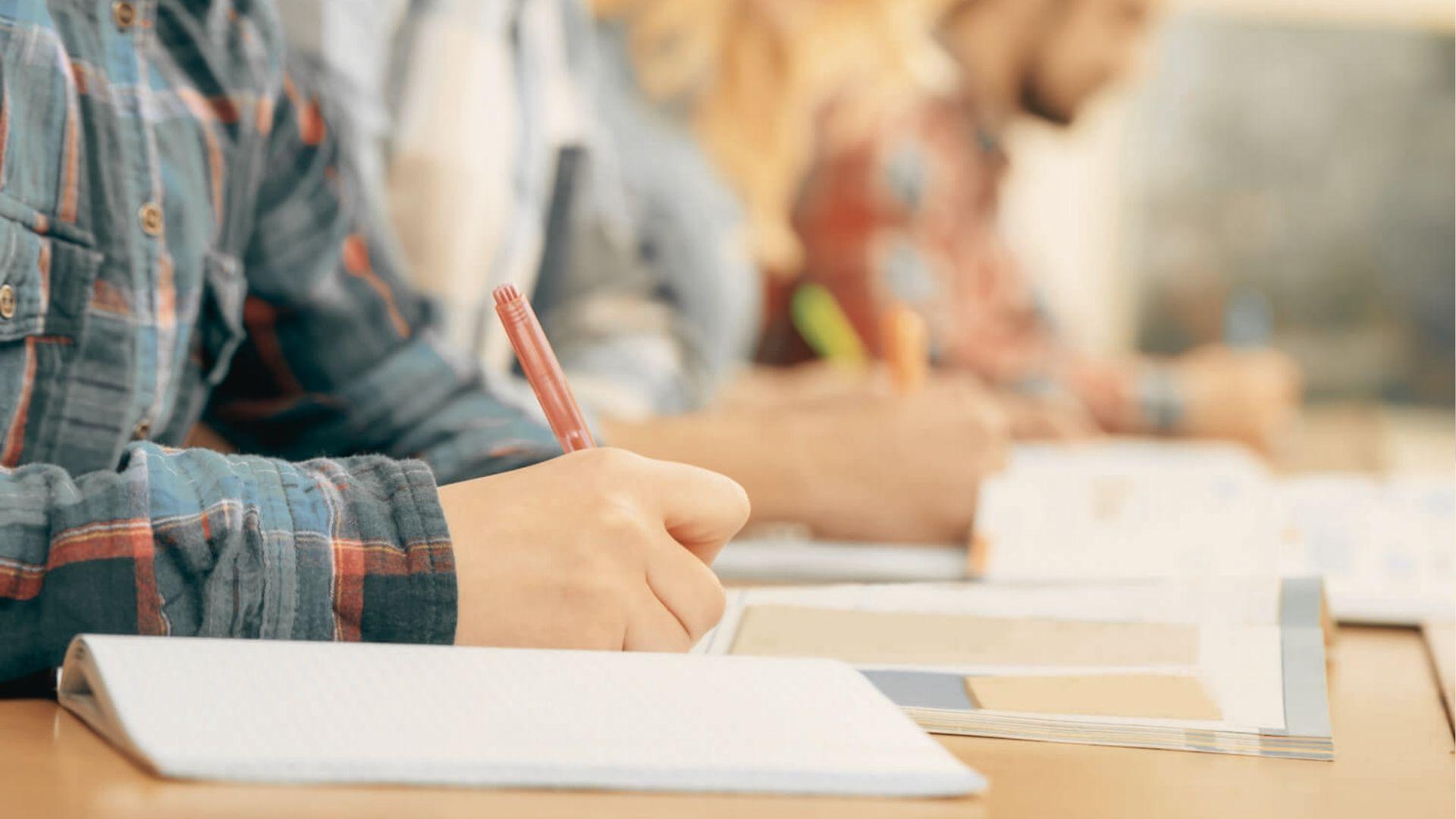 Close-up of a student's hand holding a red pen and writing in a notebook during class.