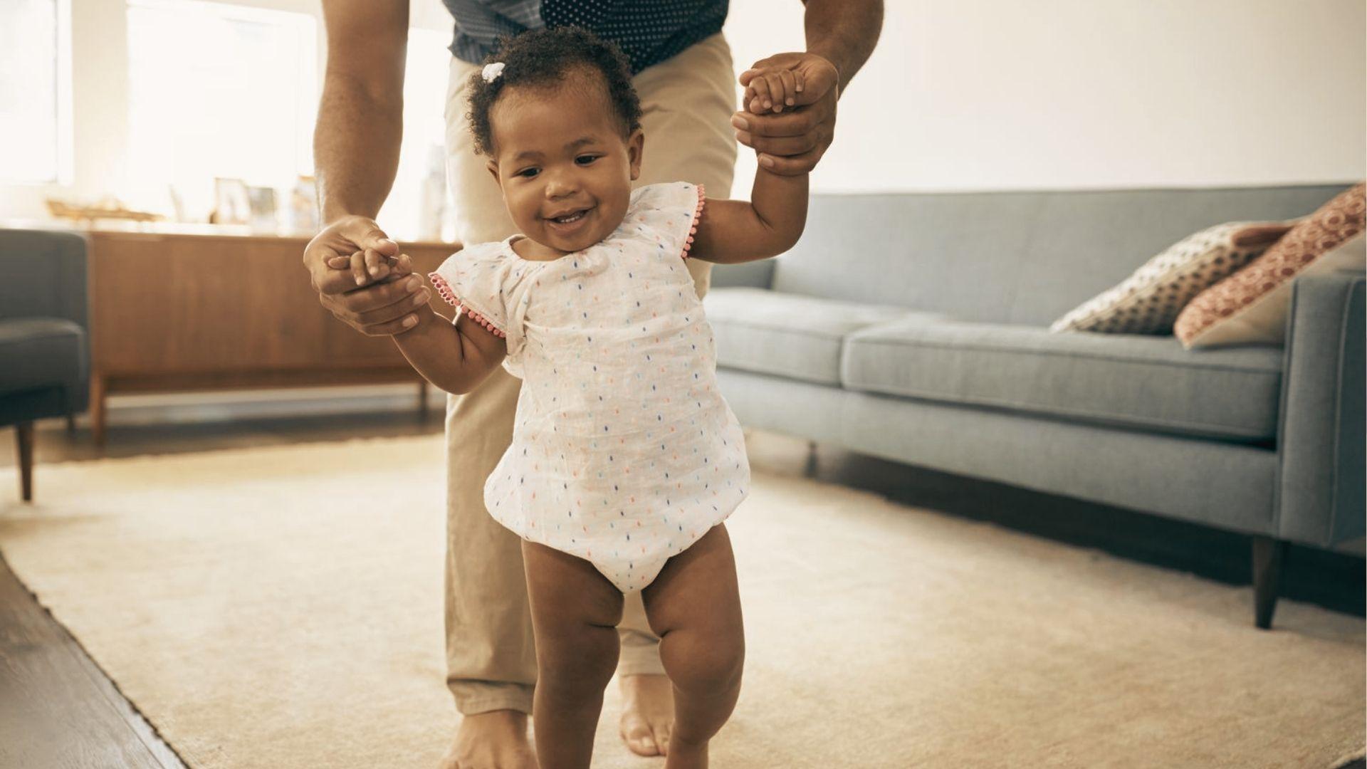 A toddler in a white romper holds an adult’s hands while practicing walking across a living room rug.