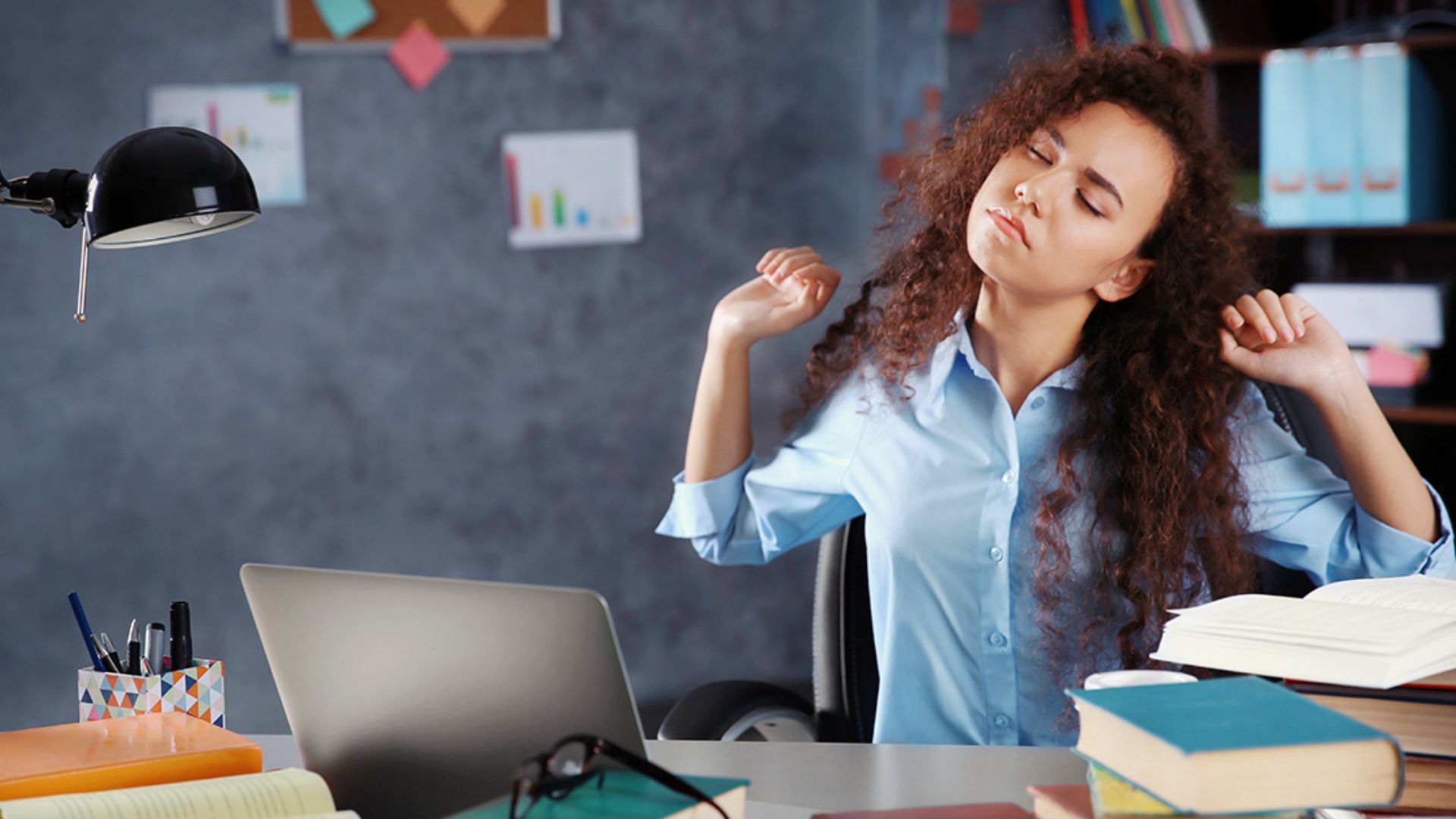 A student with curly hair stretches at her desk surrounded by books, a laptop, and a lamp.