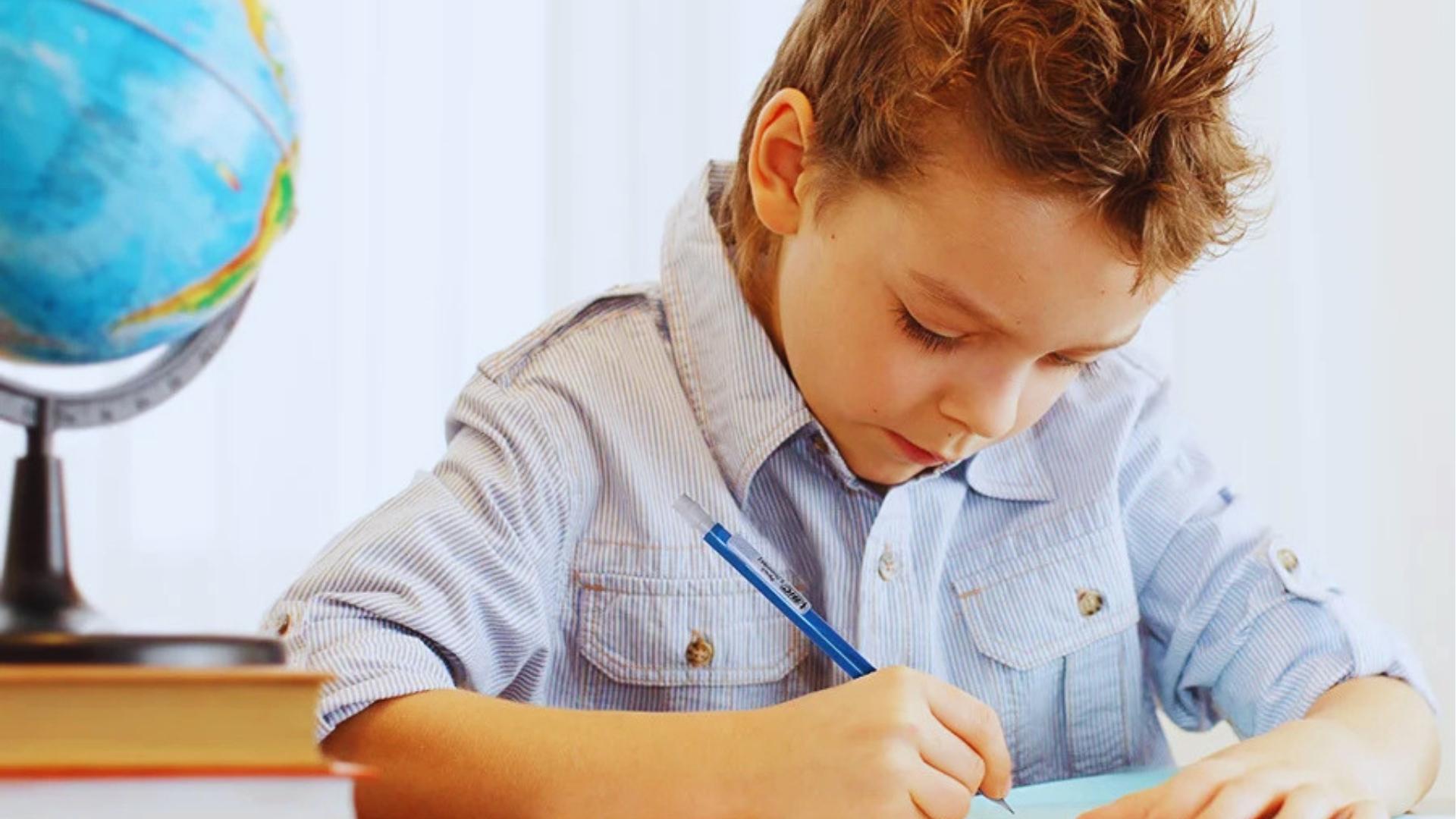 Young boy in striped shirt writing with blue pen at desk beside globe and stacked books.