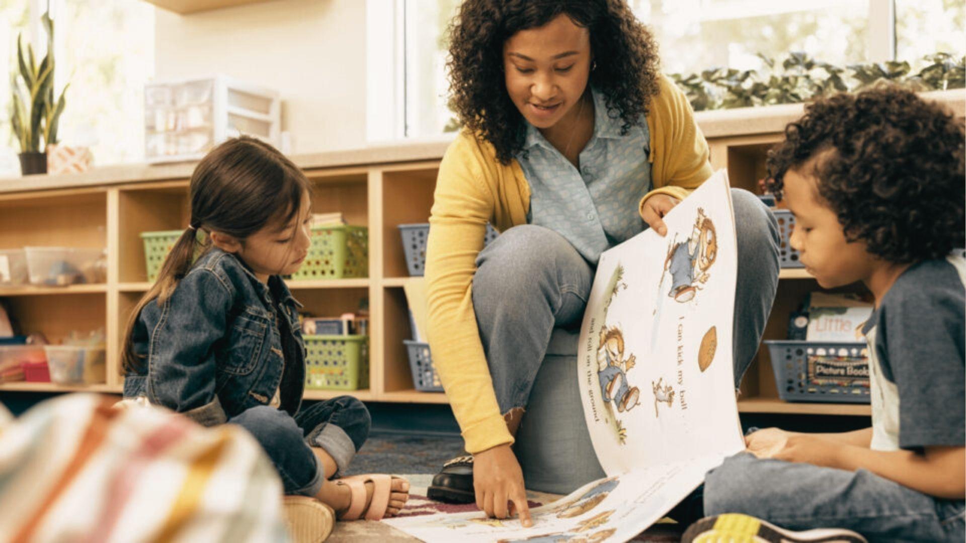 A teacher points to a large picture book while sitting on the floor with two young students in a reading nook.
