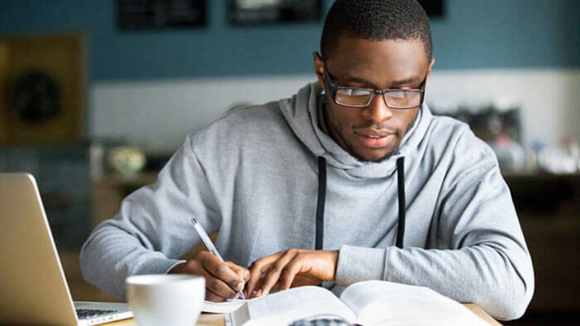 A focused student in glasses takes notes from a textbook with a laptop nearby.