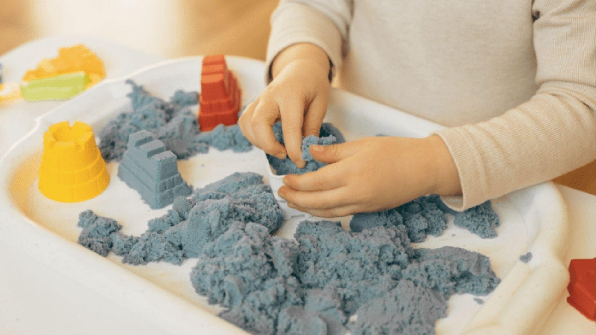 A preschooler presses blue kinetic sand into molds using their hands, building small castle shapes on a tray.