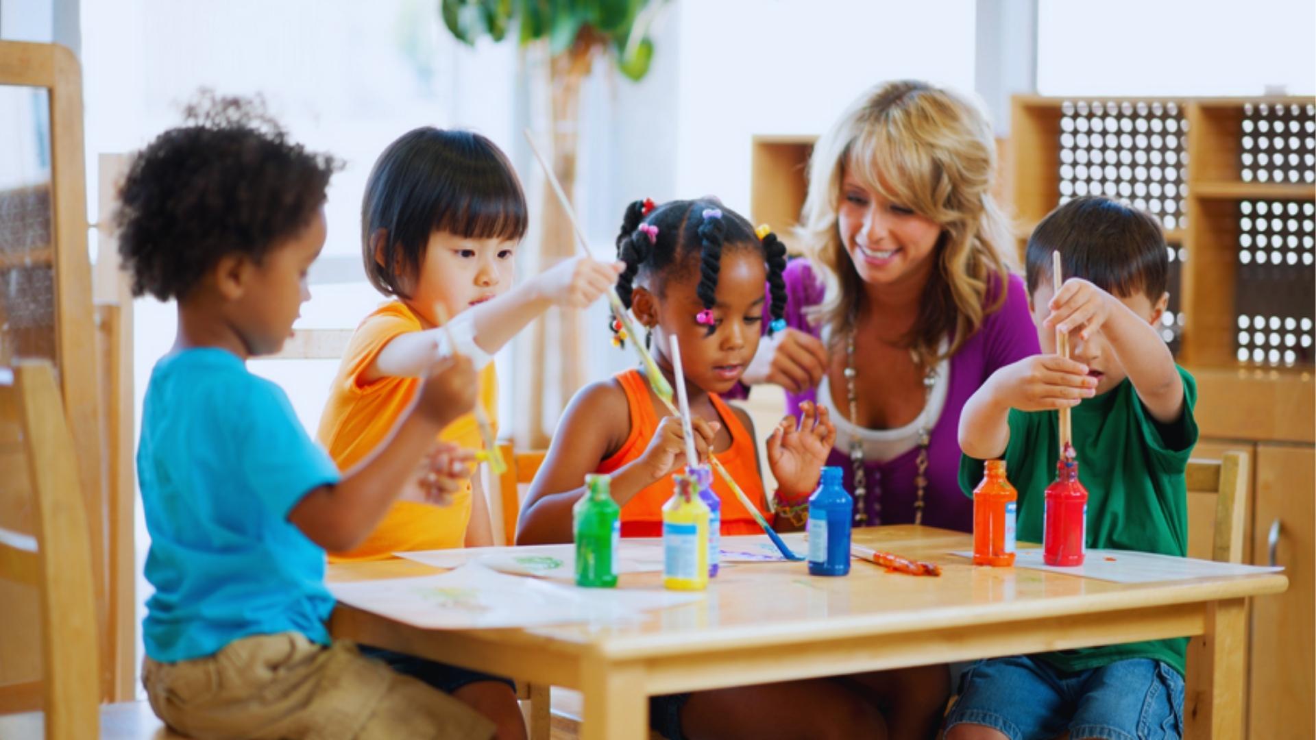Preschool teacher in purple top smiling while watching four kids paint with brushes at table with paint.