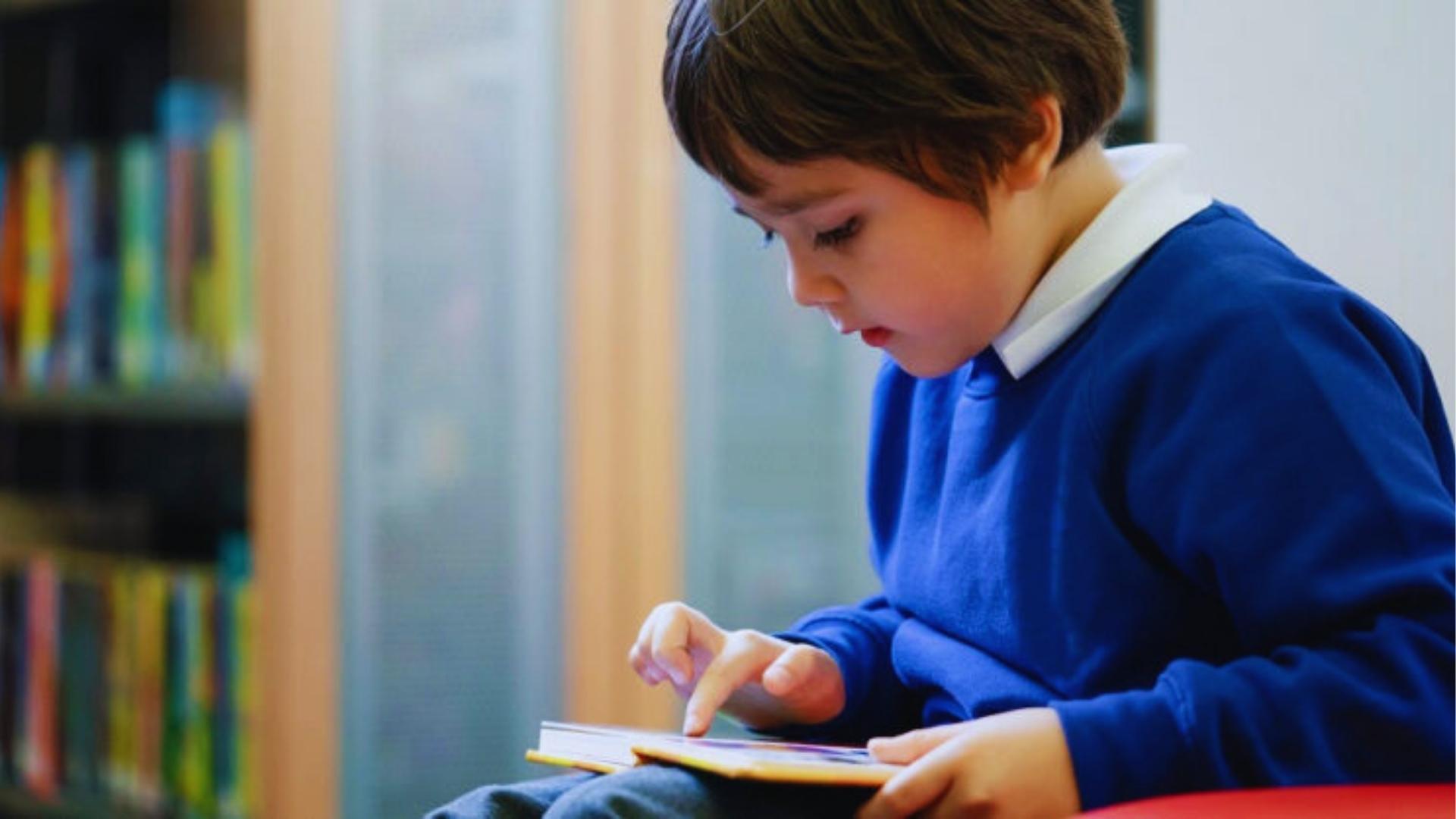Young boy in blue school sweater sitting in library pointing at book while sounding out words with focus.