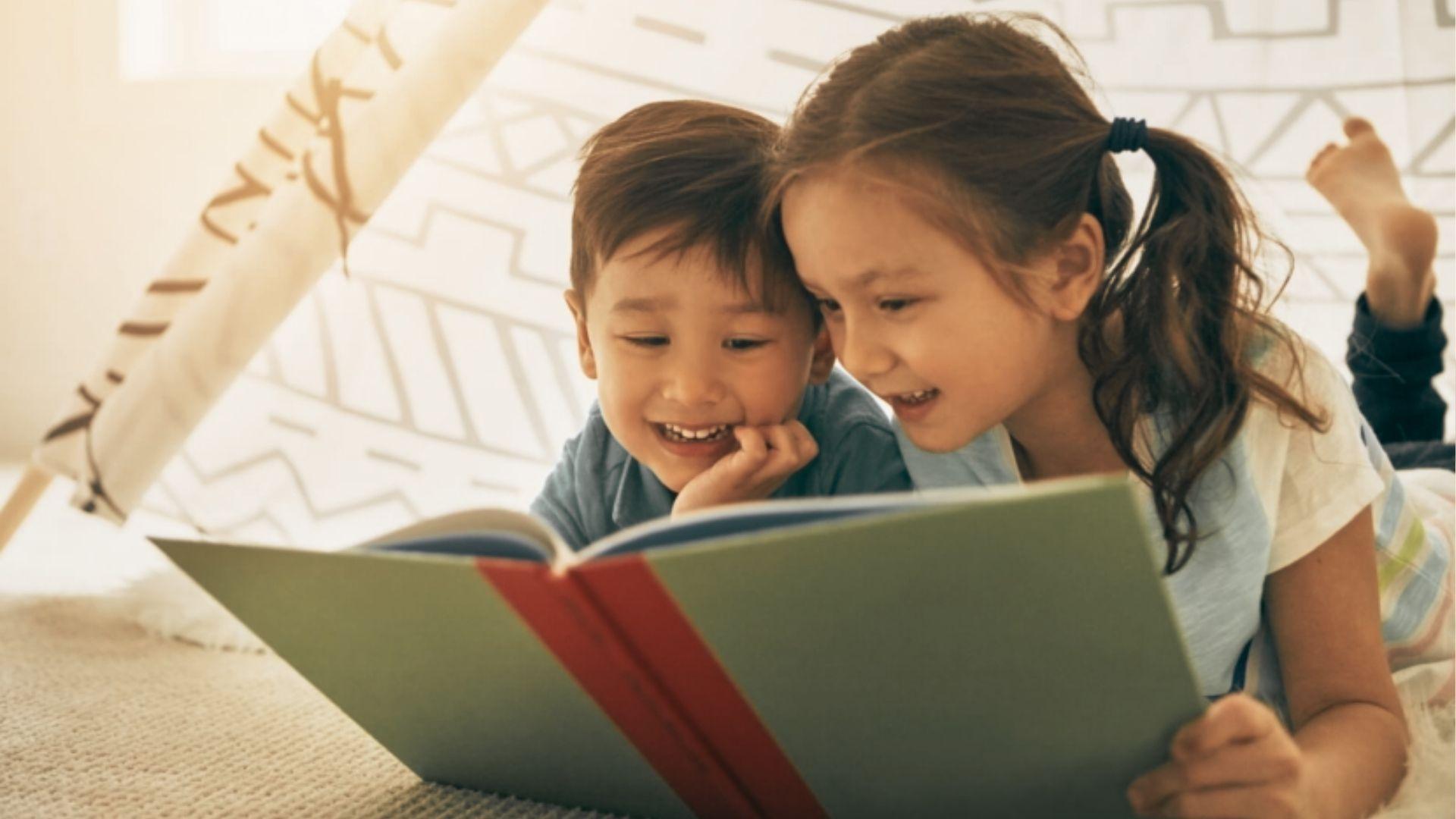 Two children lie side by side in a play tent, smiling as they share a picture book together.