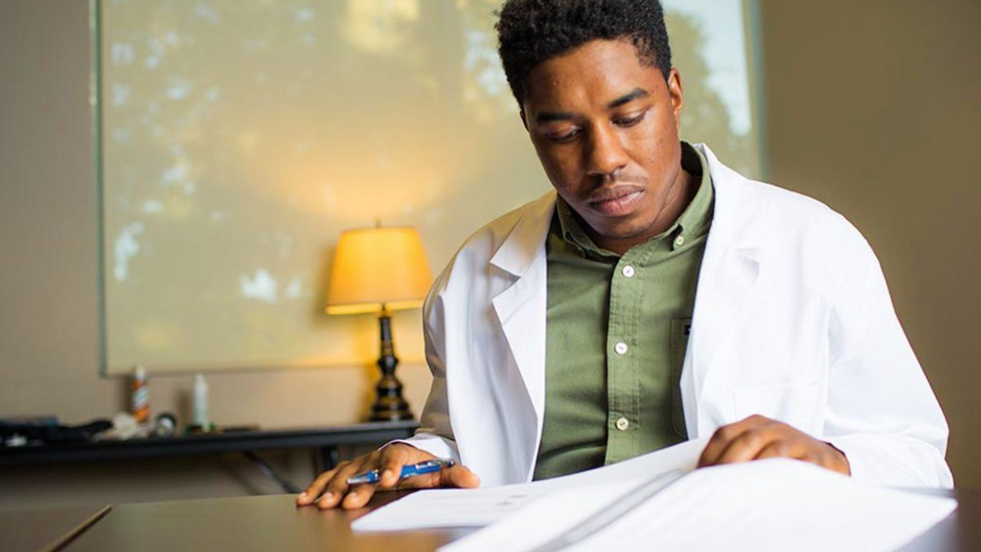 A student in a white lab coat reviews papers intently at a desk with warm lighting.