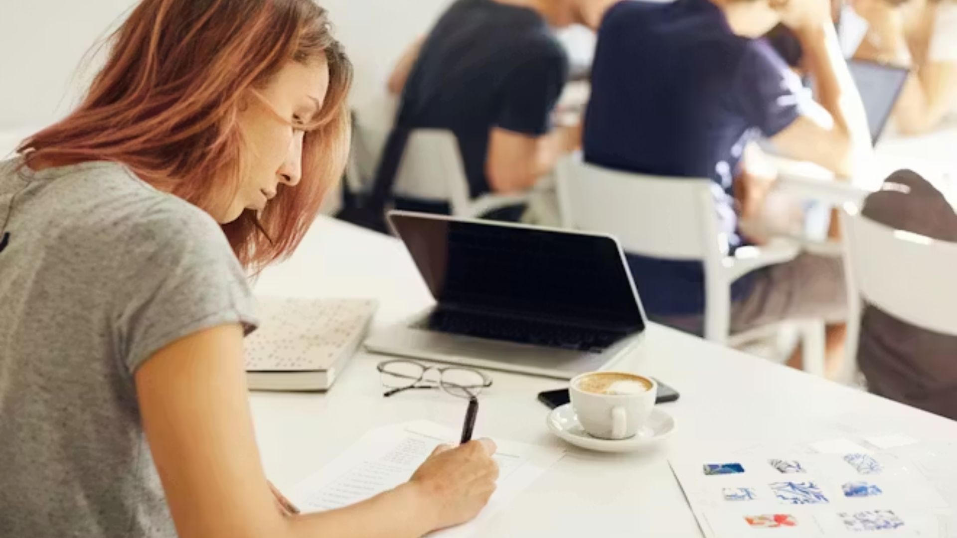 A focused student writes notes in a busy cafe with a laptop, coffee, and study materials spread out.