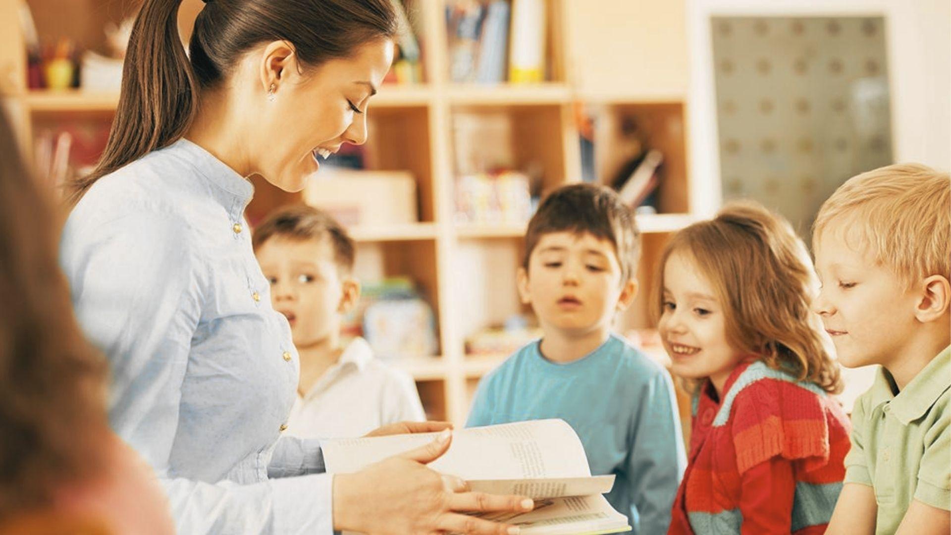 A teacher smiles while reading a book aloud to a small group of attentive preschoolers in a classroom.