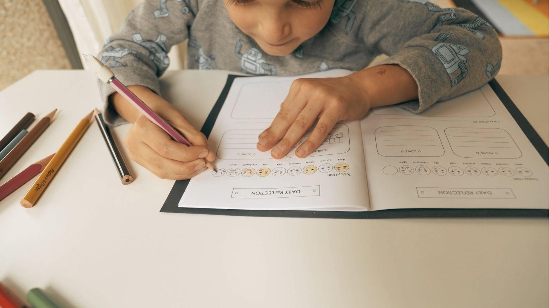 A child in a robot-print sweatshirt colors emotion icons on a "Daily Reflection" worksheet with pencils nearby.