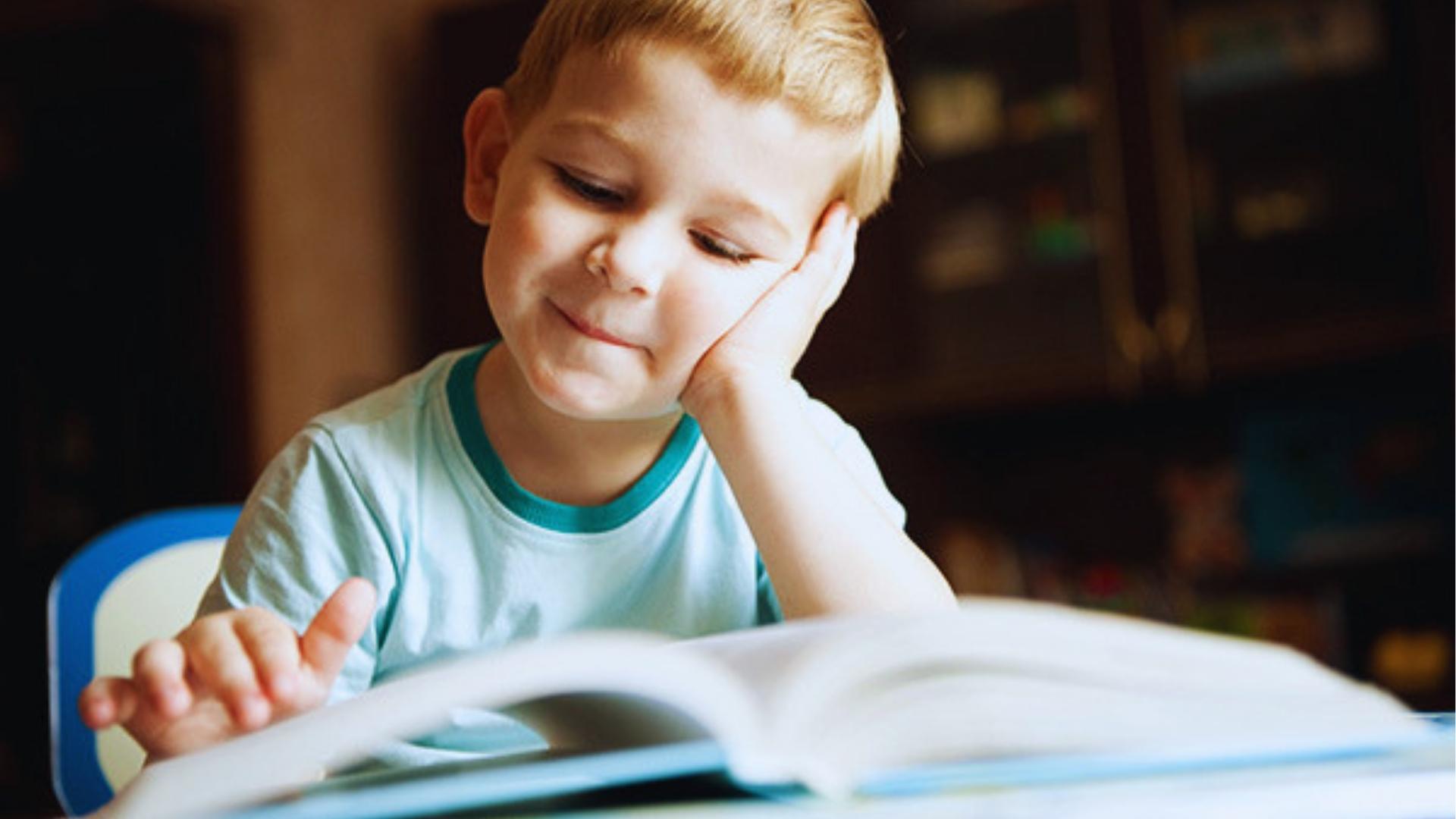 Smiling blonde boy in teal shirt resting head on hand while reading an open book at home desk.
