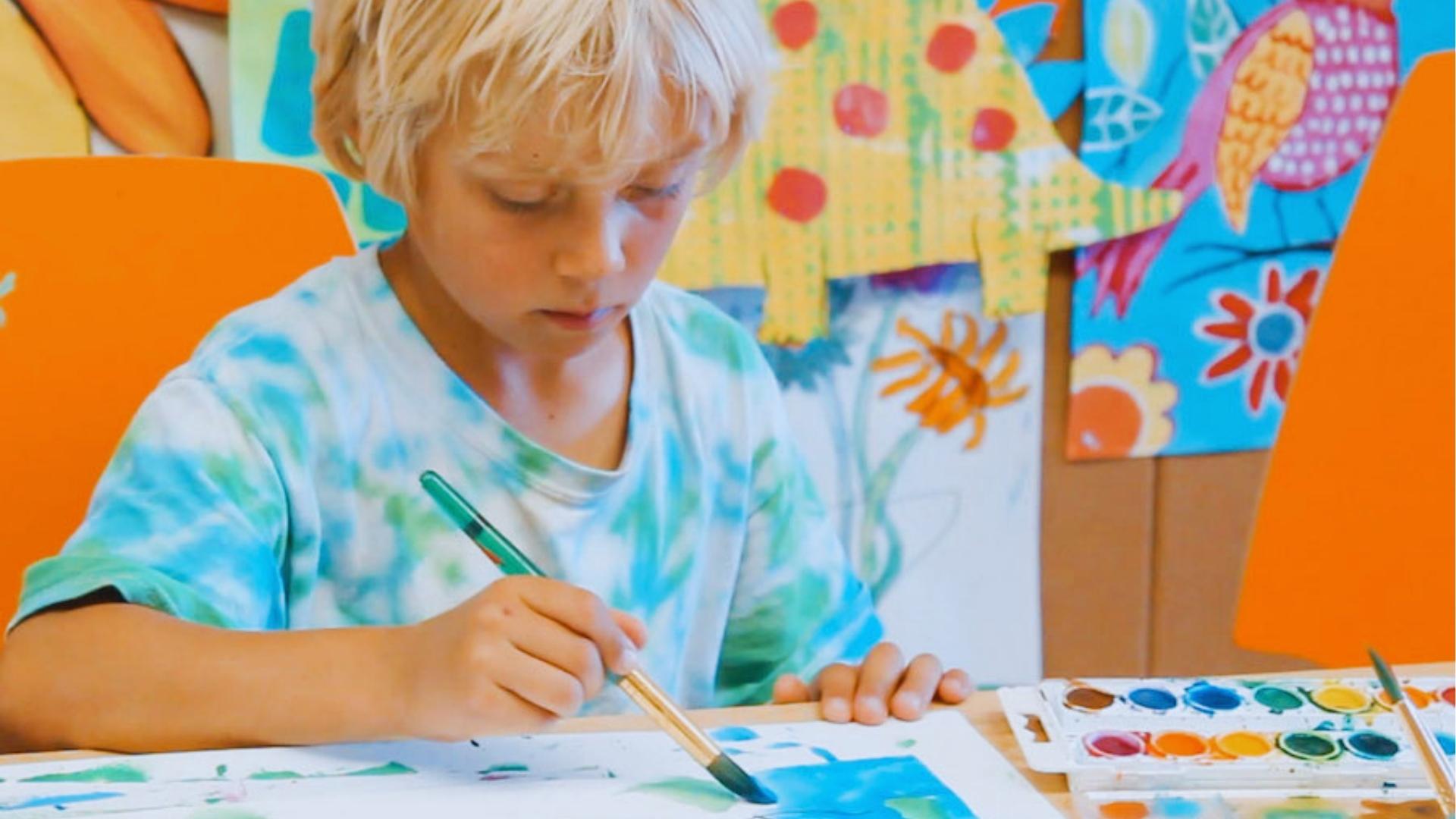 Blonde child in tie-dye shirt focused on watercolor painting with brush at desk beside colorful classroom artwork.