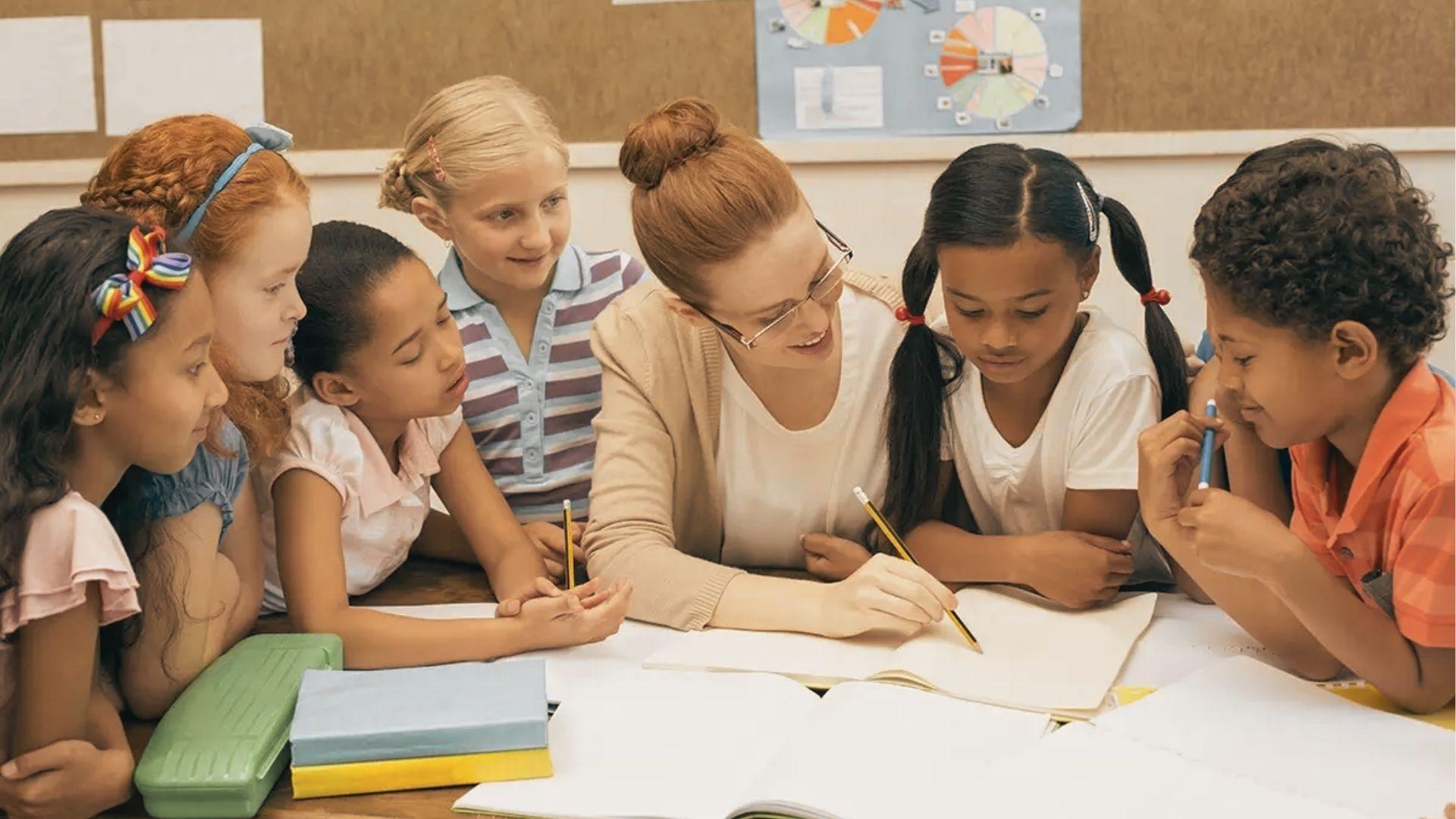 Teacher leans over a table helping six diverse students who are writing and reading together.