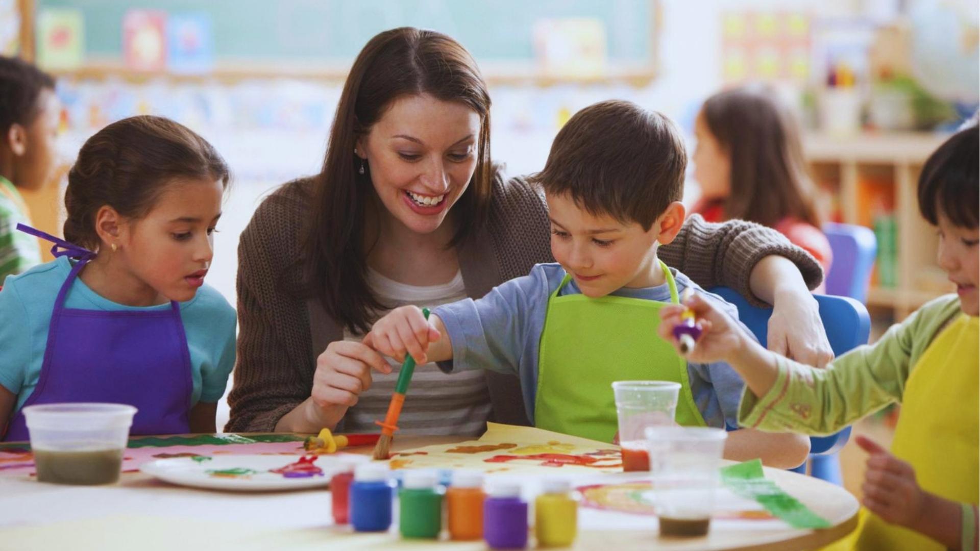 Smiling teacher helping kids in colorful aprons paint together at classroom table with brushes and paint cups.