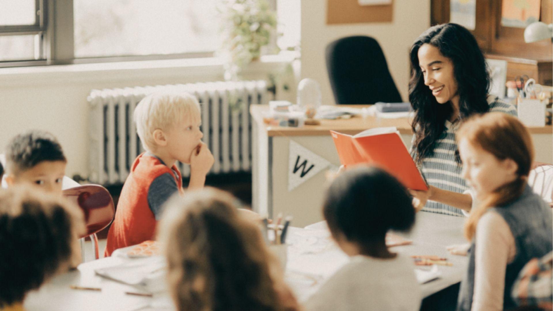 A teacher reads from a bright orange book to a circle of attentive students in a sunny classroom.