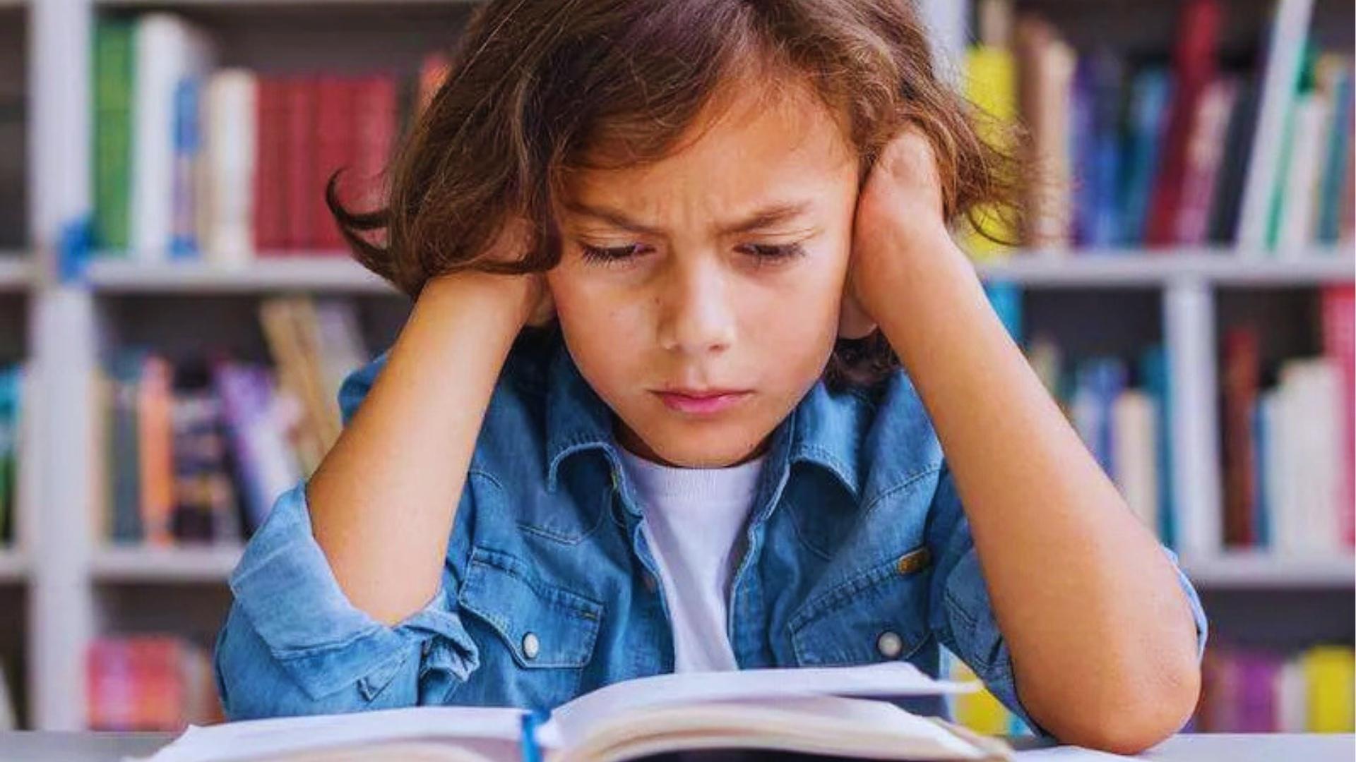 Child in denim shirt covering ears while struggling to read open book in front of bookshelf.