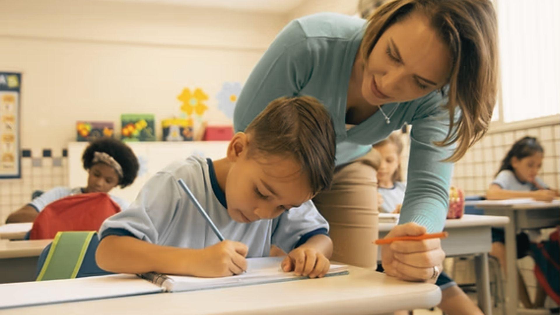 A teacher leans over a student’s desk, gently guiding him with a pencil as he writes in a classroom.