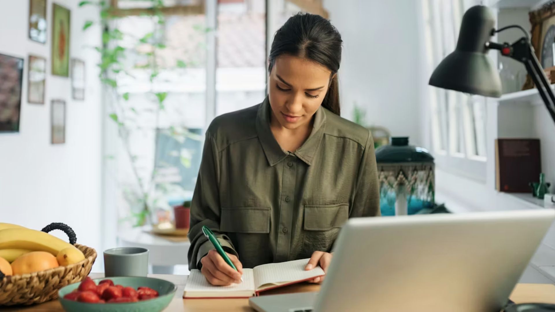A student takes notes in a notebook at a bright home desk with a laptop and fruit nearby.