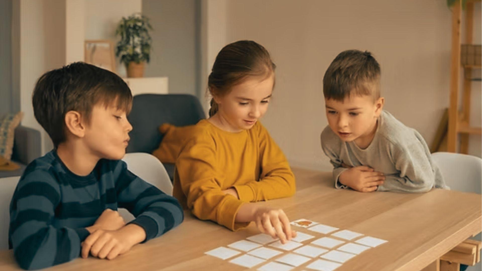 Three children sit at a wooden table playing a matching card game, focused on rows of face-down cards.