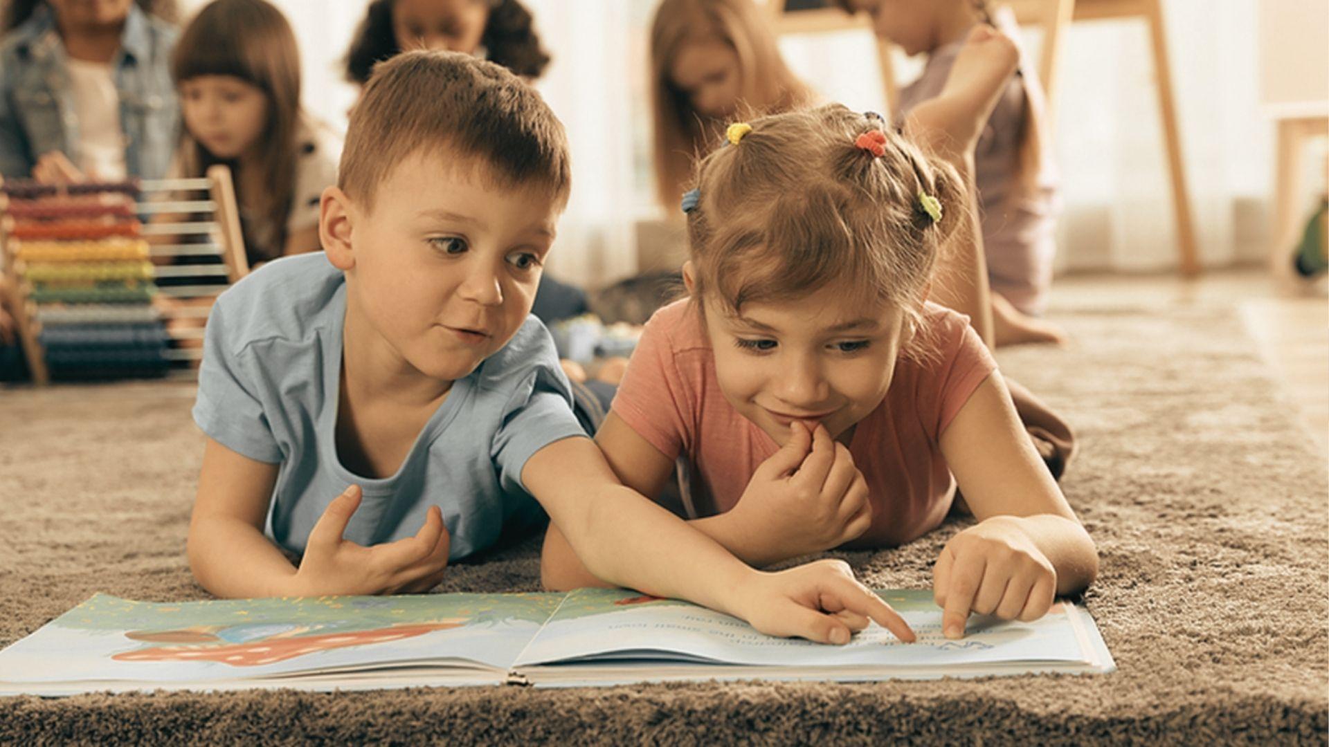 Two children lie on a carpet pointing at words in a large picture book, with classmates in the background.