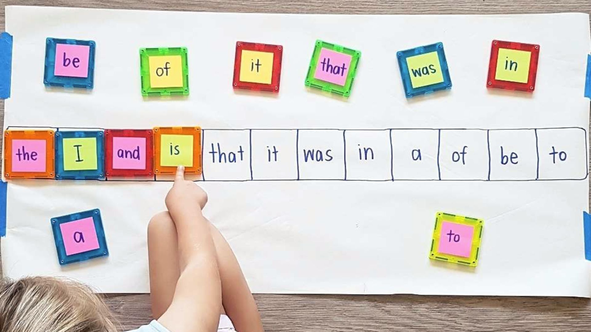 A child matches colorful magnetic tiles with sight words to a handwritten word chart on paper.