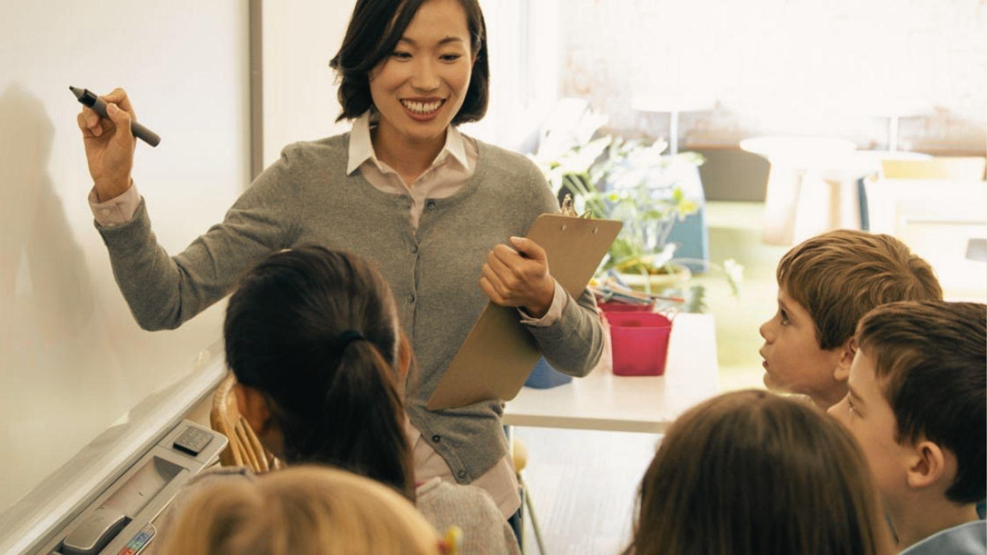 A teacher smiles while writing on a whiteboard as students watch attentively from their desks.