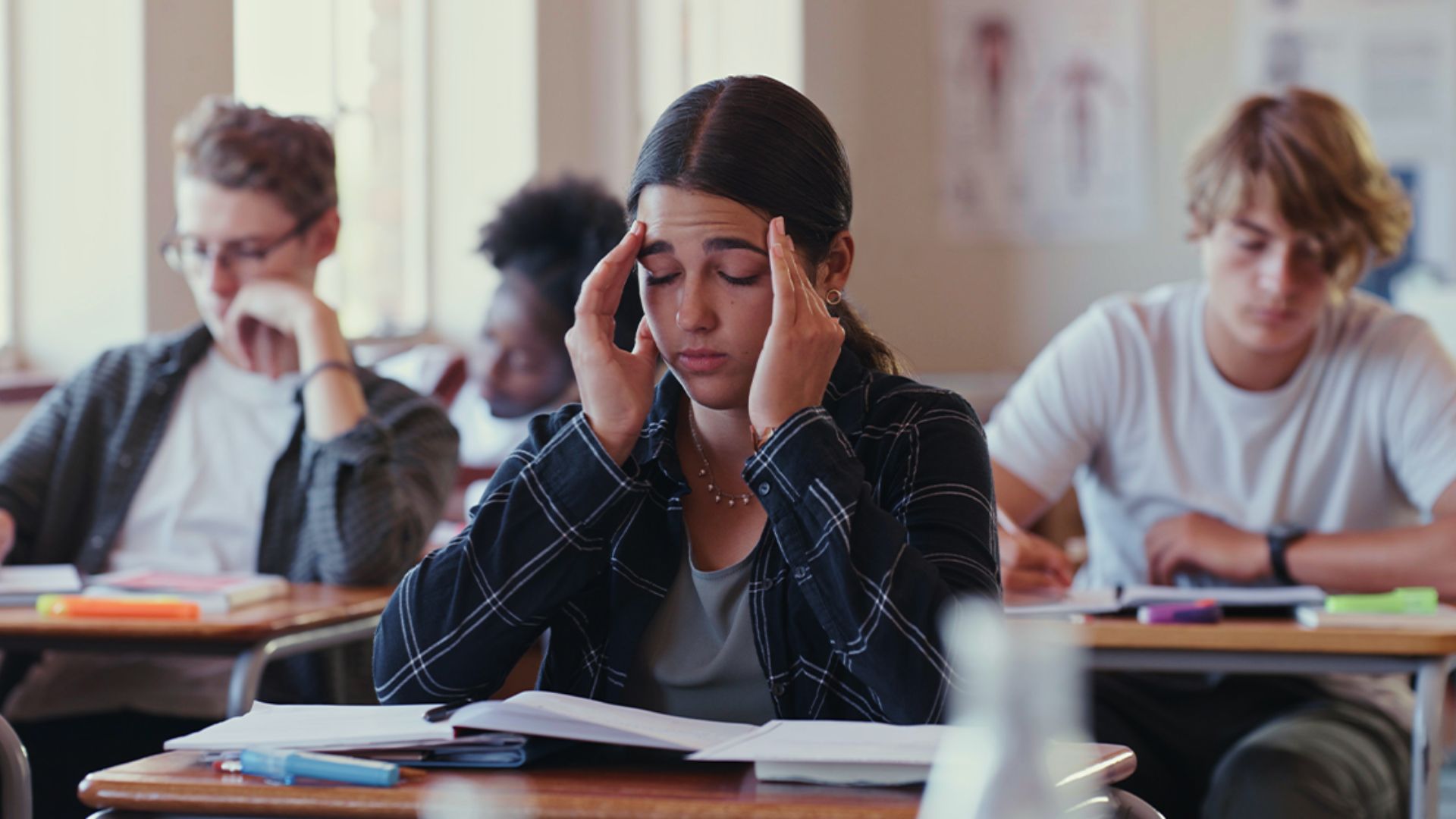 A student holds her head in stress while studying with classmates in a classroom.